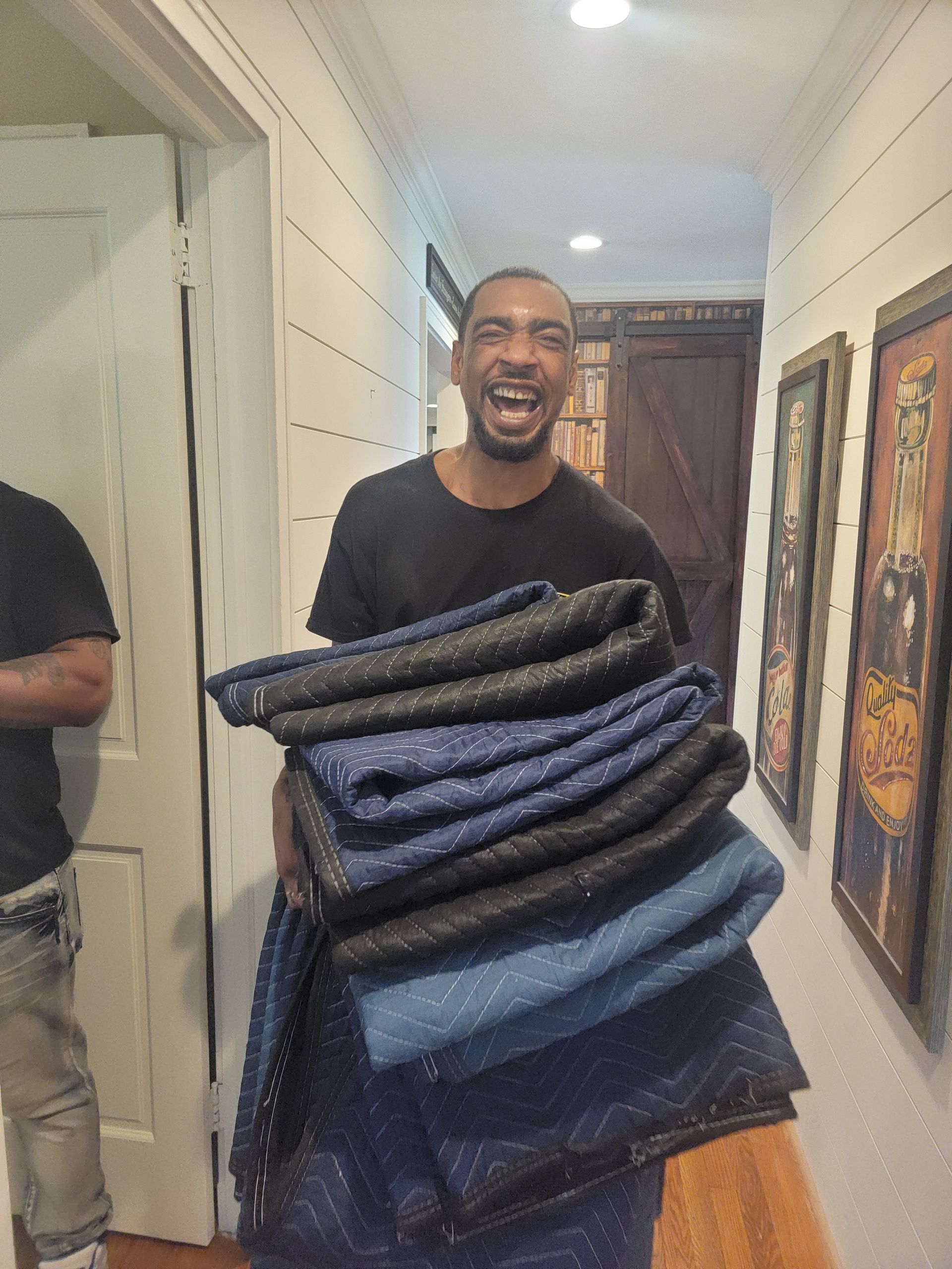 Smiling man holding a stack of blue and black moving blankets in a hallway. He is wearing a black shirt.