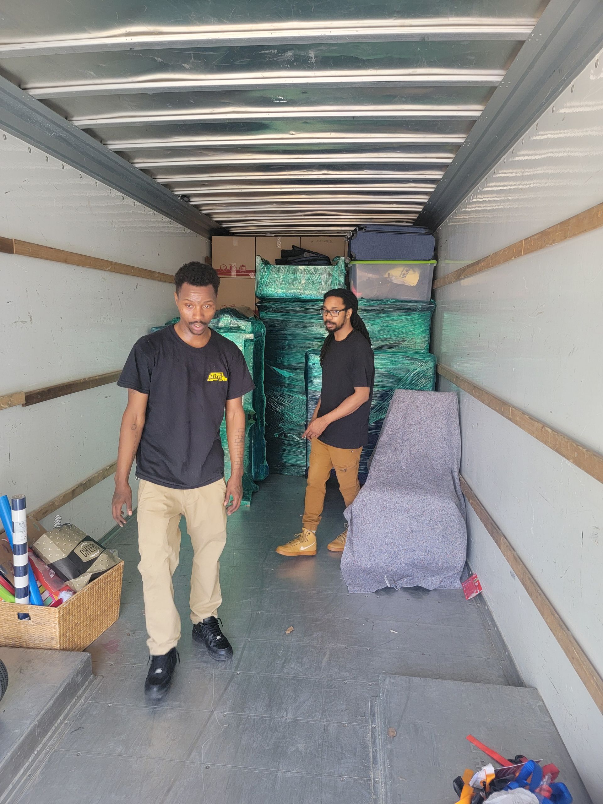 Two movers stand in an empty truck, boxes and wrapped items stacked behind them. One wears tan pants, the other khaki.