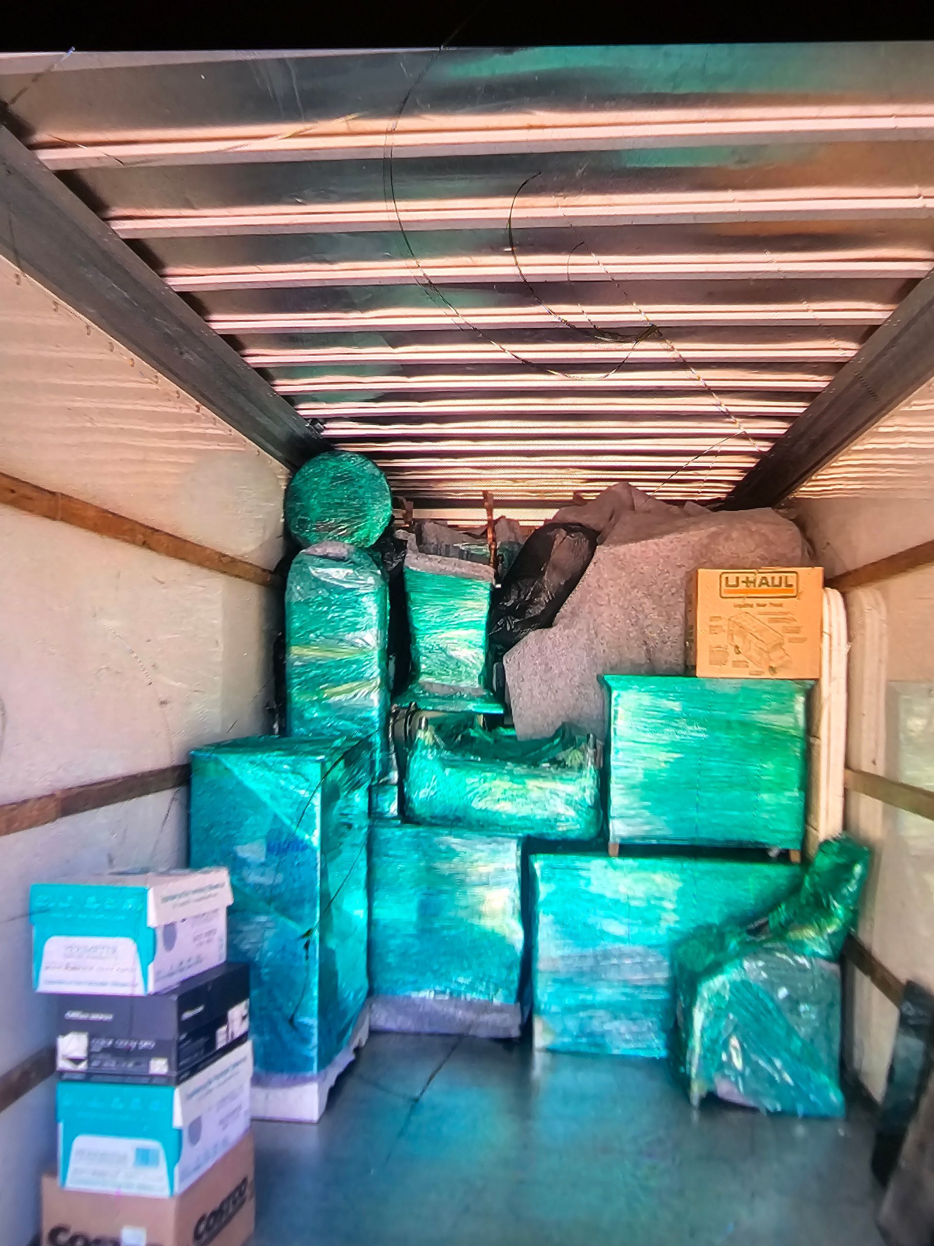 Inside a moving truck, various items wrapped in green plastic are stacked. Boxes and other contents are tightly packed for transport.