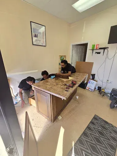 Three people working on a brown desk in a room with light-colored walls and flooring.