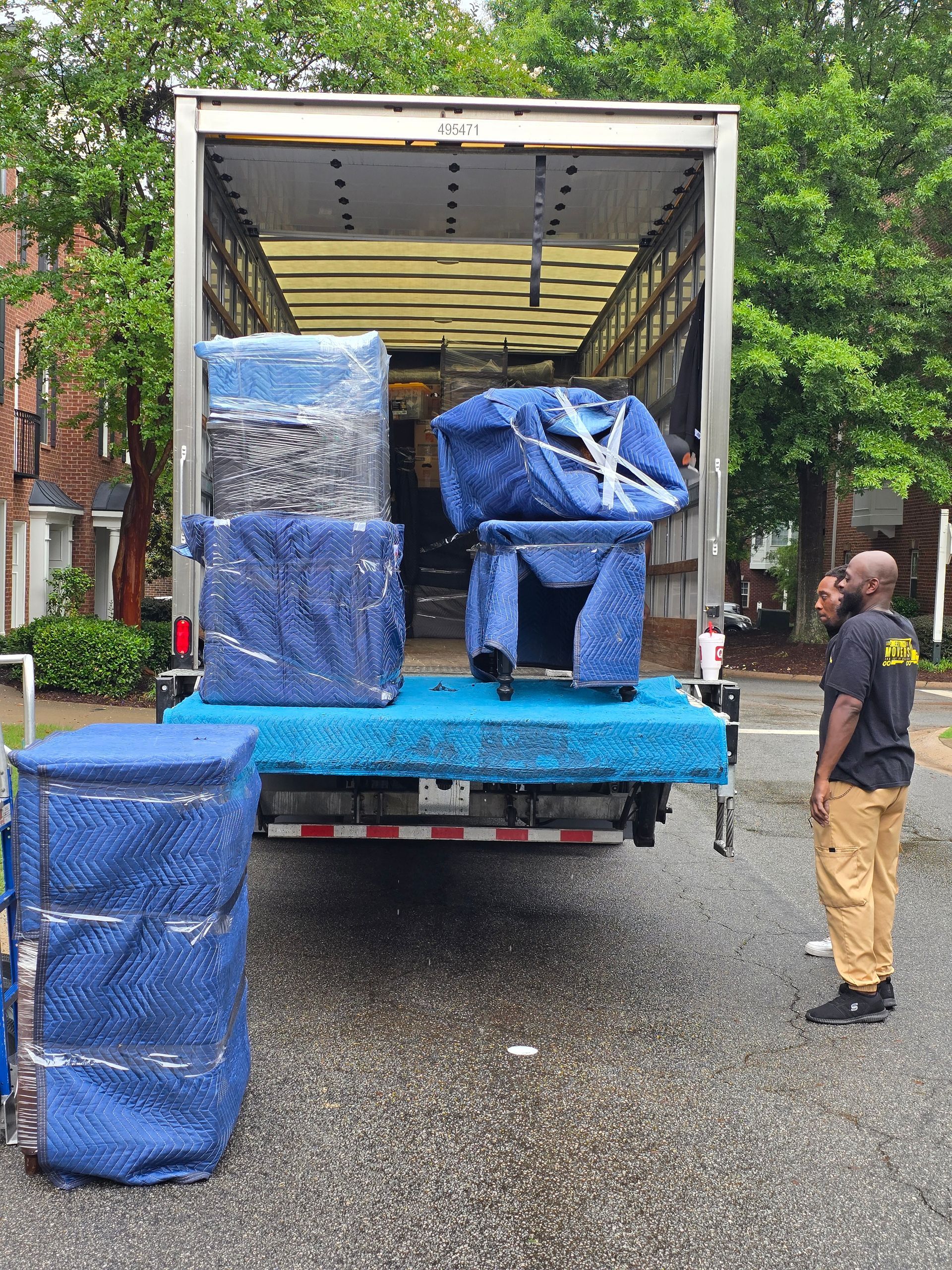 A moving truck being loaded with furniture wrapped in blue bubble wrap. A man in a black shirt and khaki pants watches.