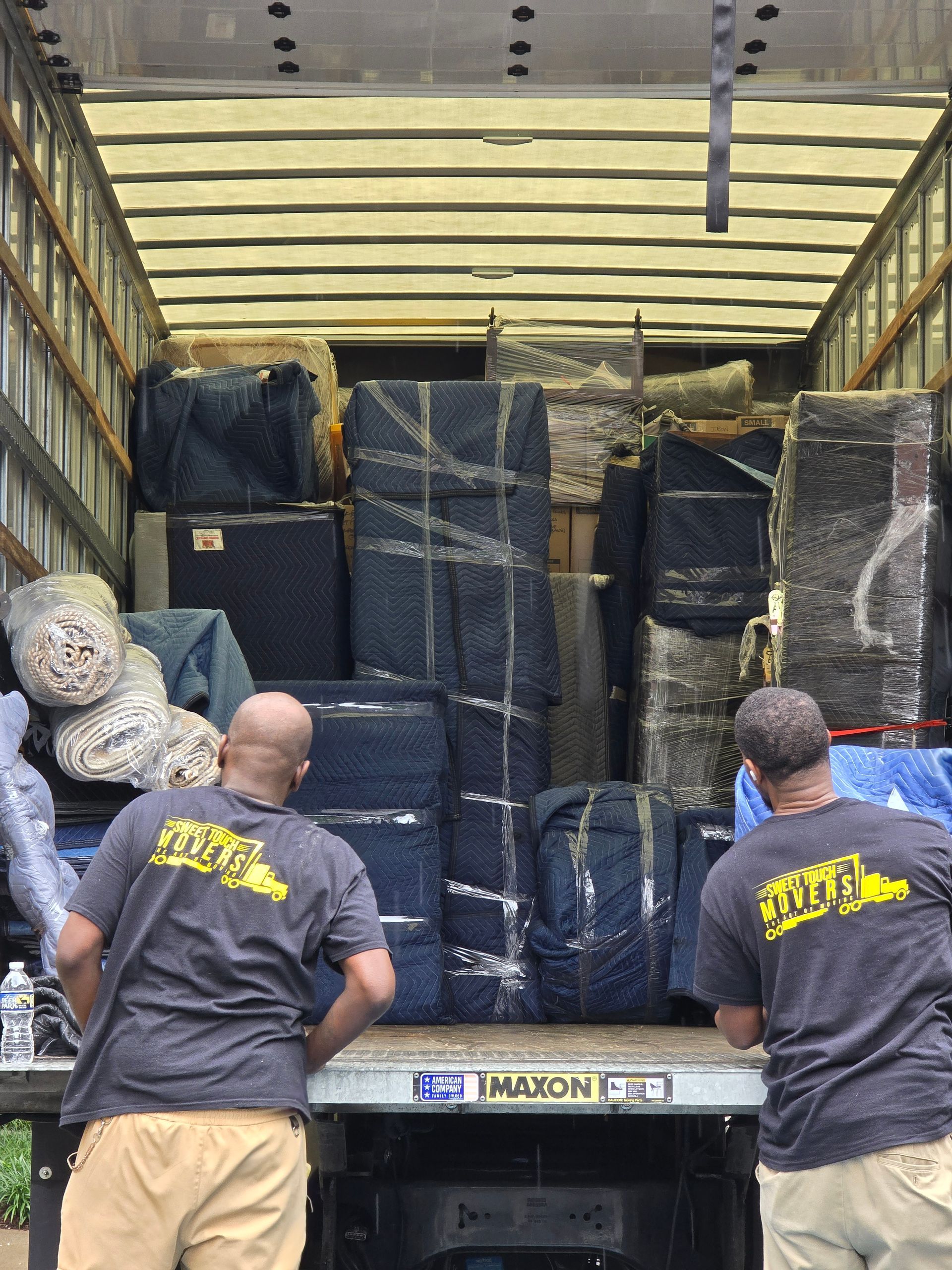 Two movers loading a packed moving truck with furniture and wrapped items. They wear grey shirts with a company logo.