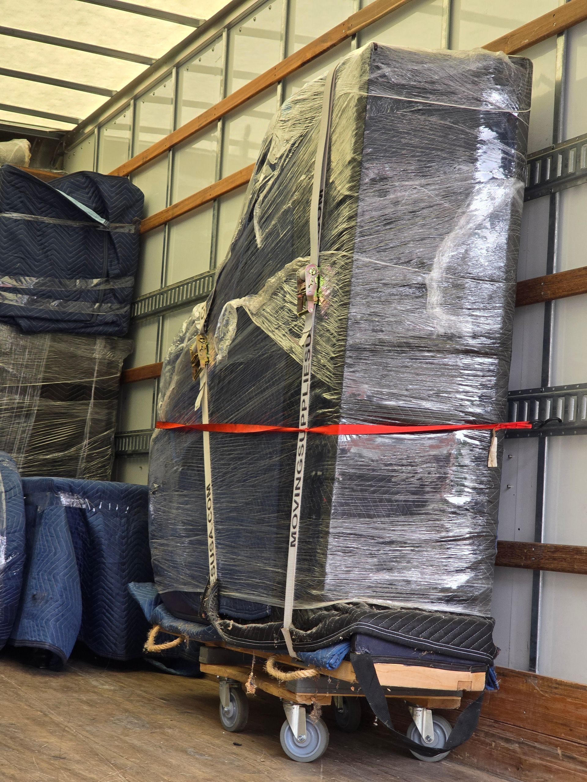 Piano, wrapped in black blankets and plastic, secured with straps on a dolly inside a moving truck.