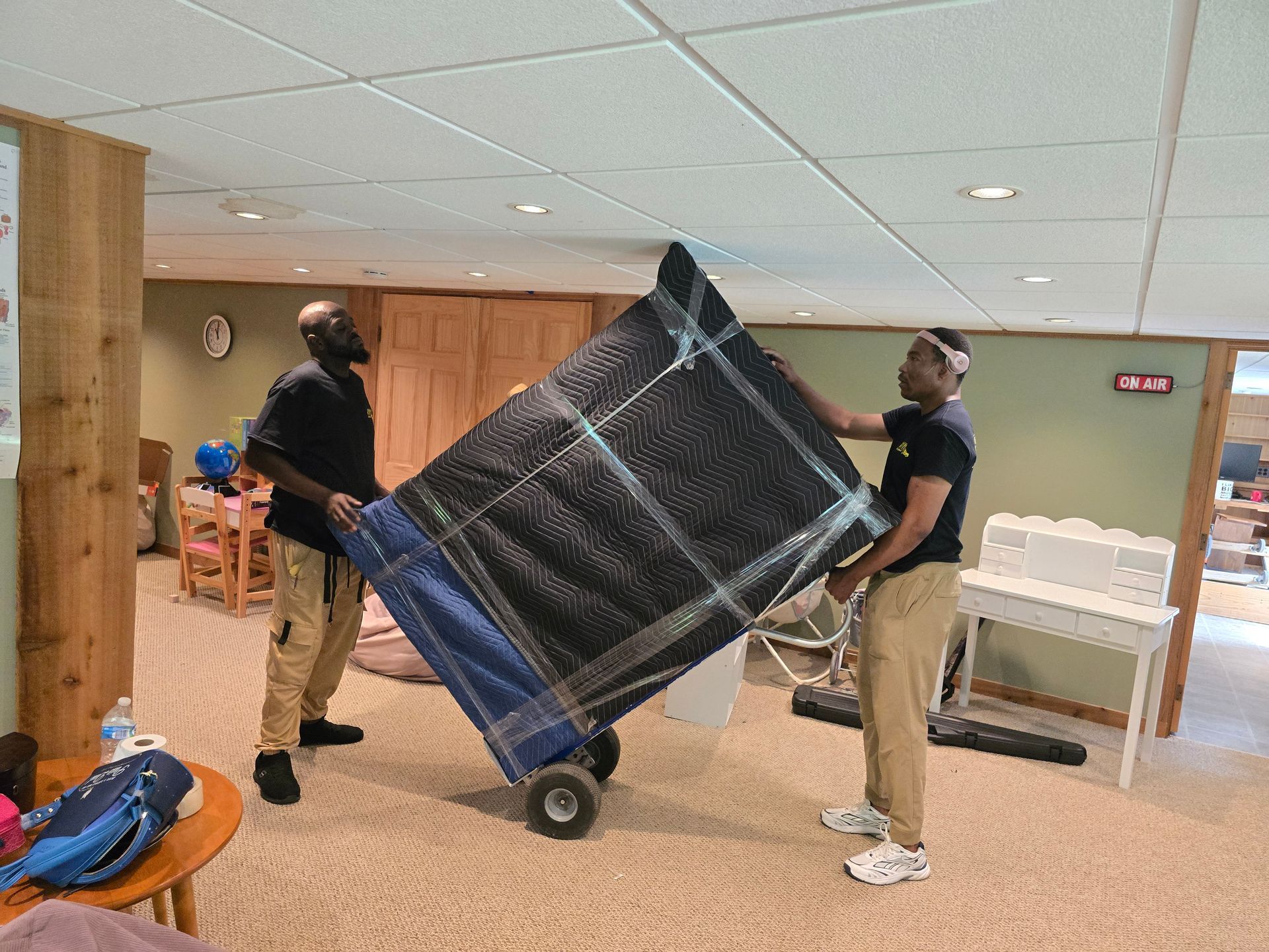 Two men lift a large, wrapped object onto a dolly in a basement. One man is on the left wearing a black shirt and khaki pants, the other on the right wearing a black shirt, khaki pants, and a white headband.