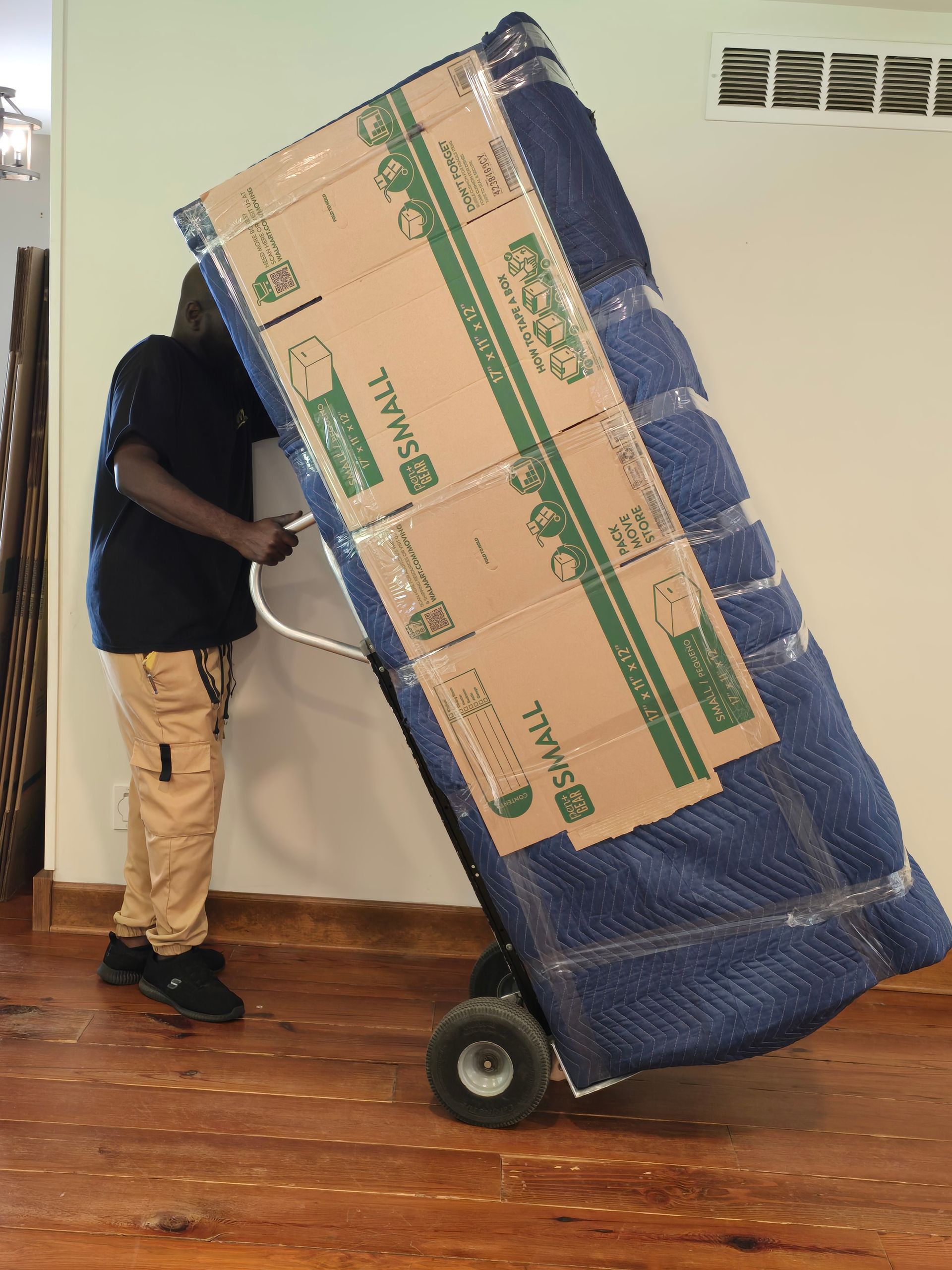 A person using a dolly to move a large, wrapped mattress. The mattress is covered in blue bubble wrap and cardboard.