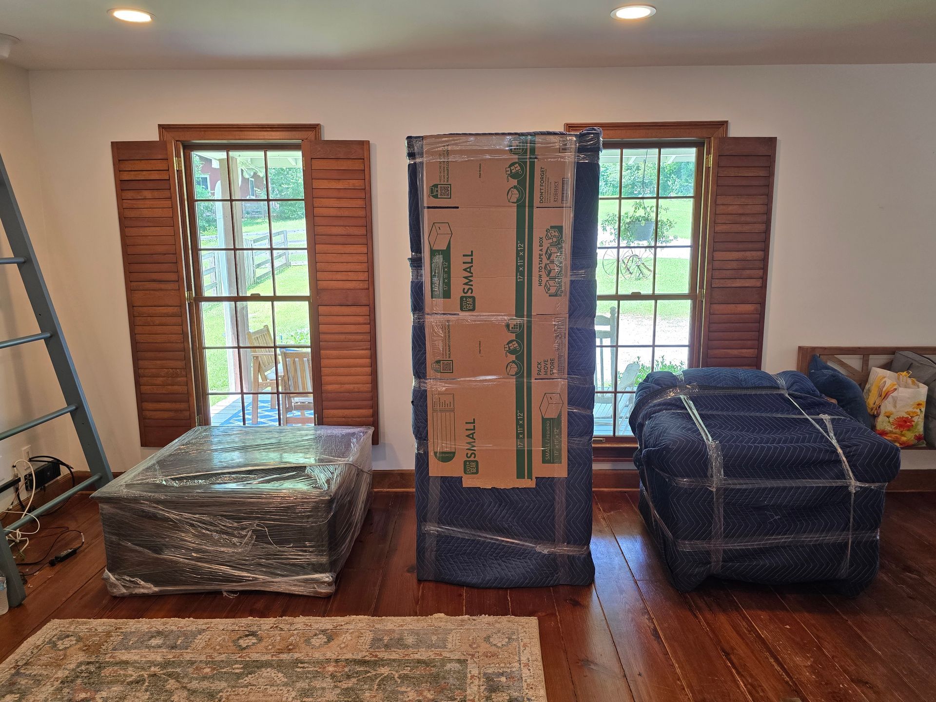 Three wrapped furniture pieces in a room with shuttered windows. A ladder leans against the wall.