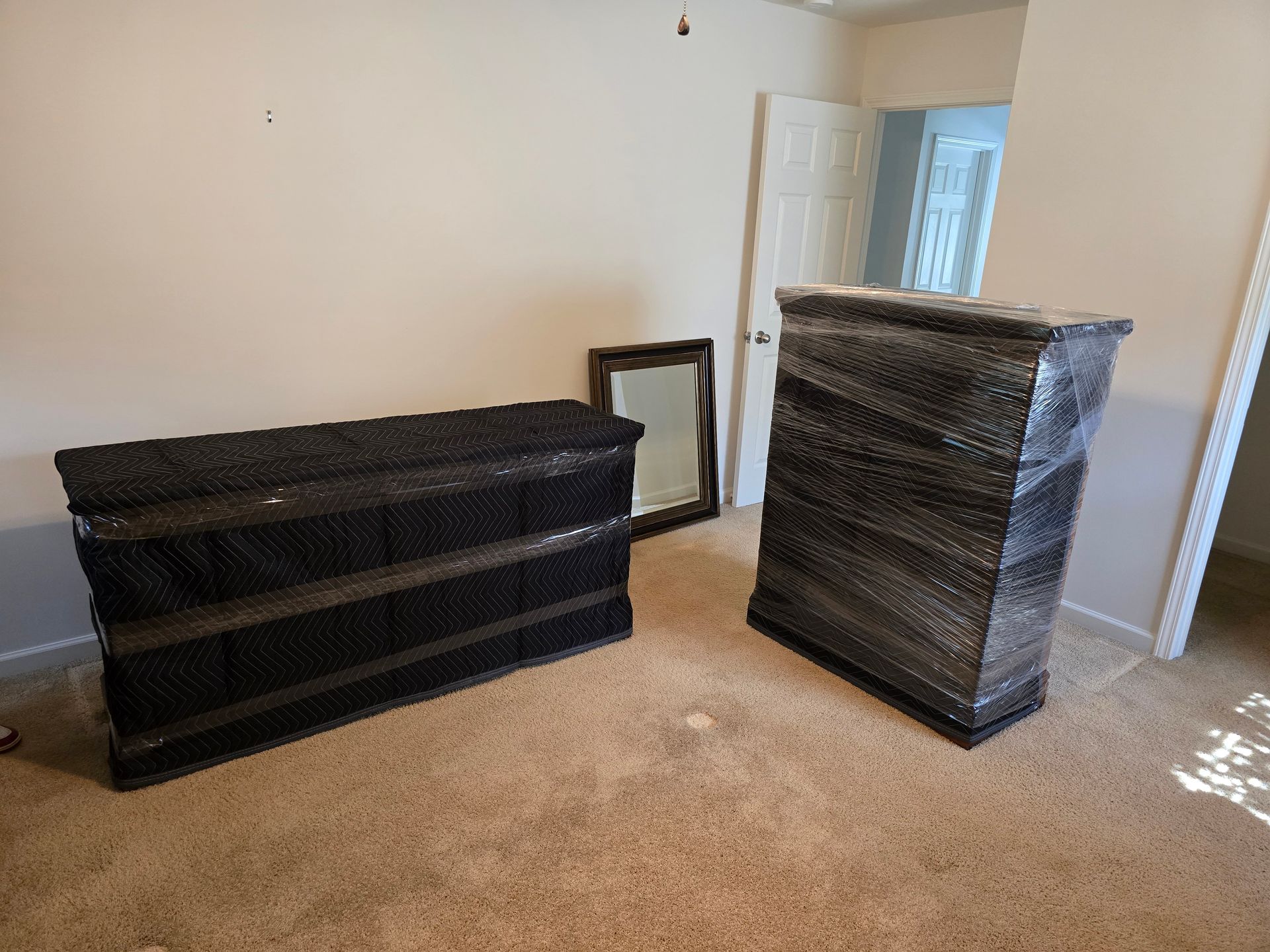 Two pieces of furniture wrapped in black plastic are in a room with beige carpet and a mirror. One piece is tall and narrow, the other is long and low.
