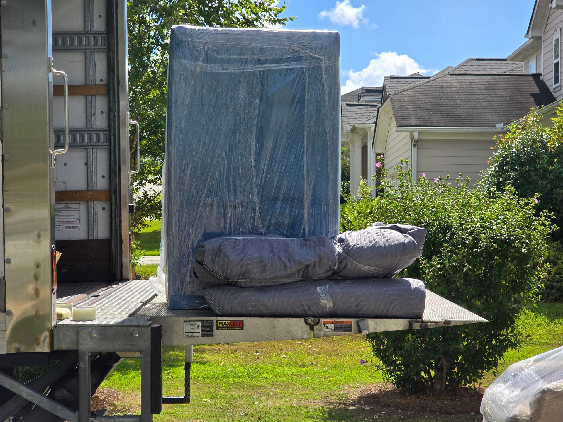A large, wrapped item and cushions are on the liftgate of a truck in a residential area. The items are wrapped in blue and gray.