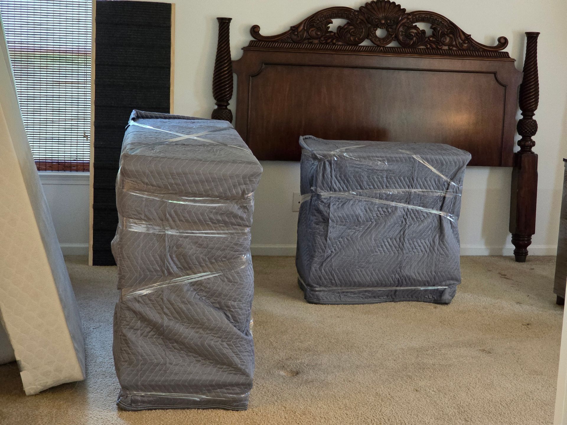 Wrapped furniture, including two nightstands and a headboard, in a bedroom with neutral carpet and a window with blinds.
