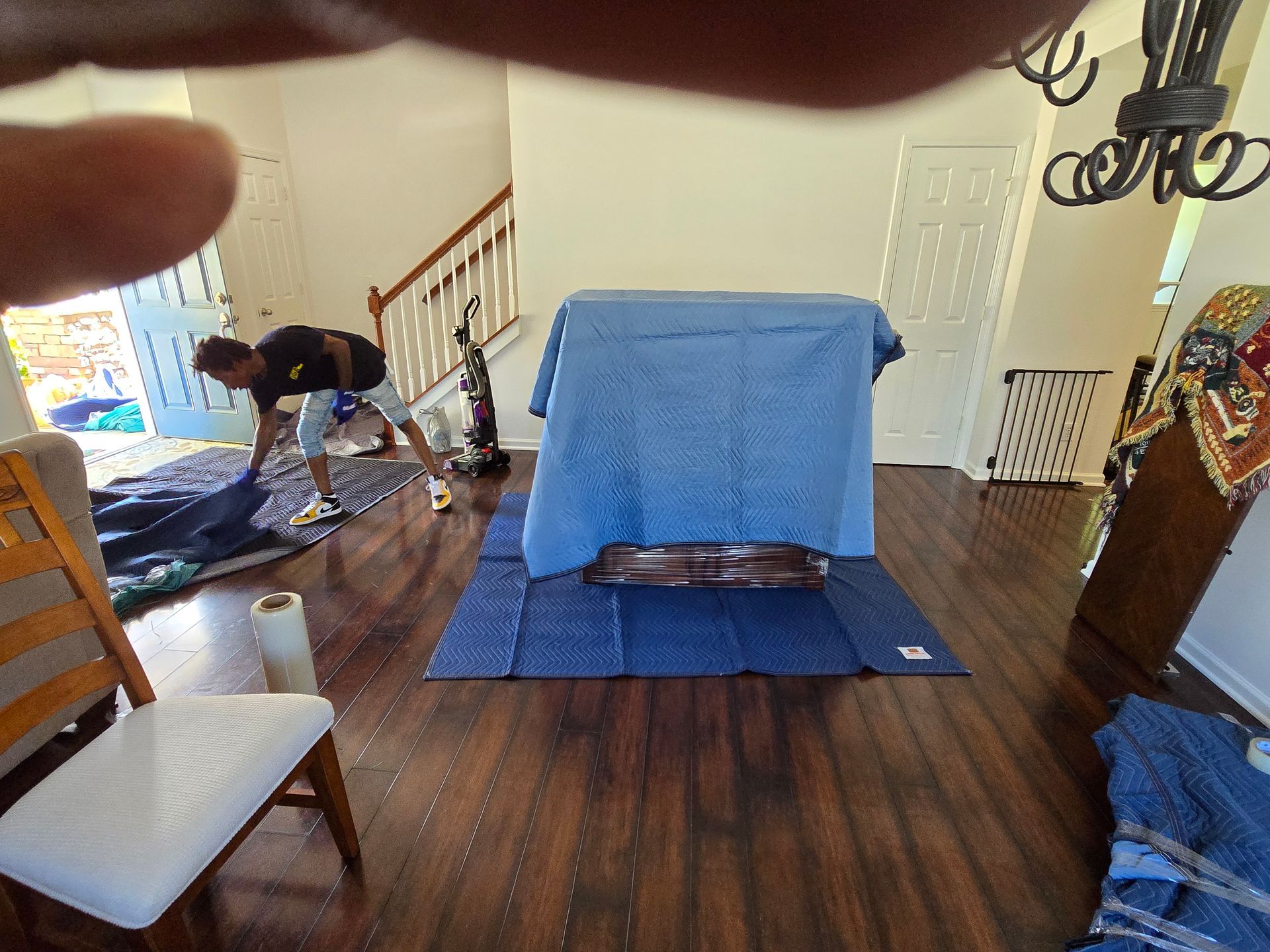 Person covering a table with blue material, another person in background. Interior, wooden floors, and open doorway visible.