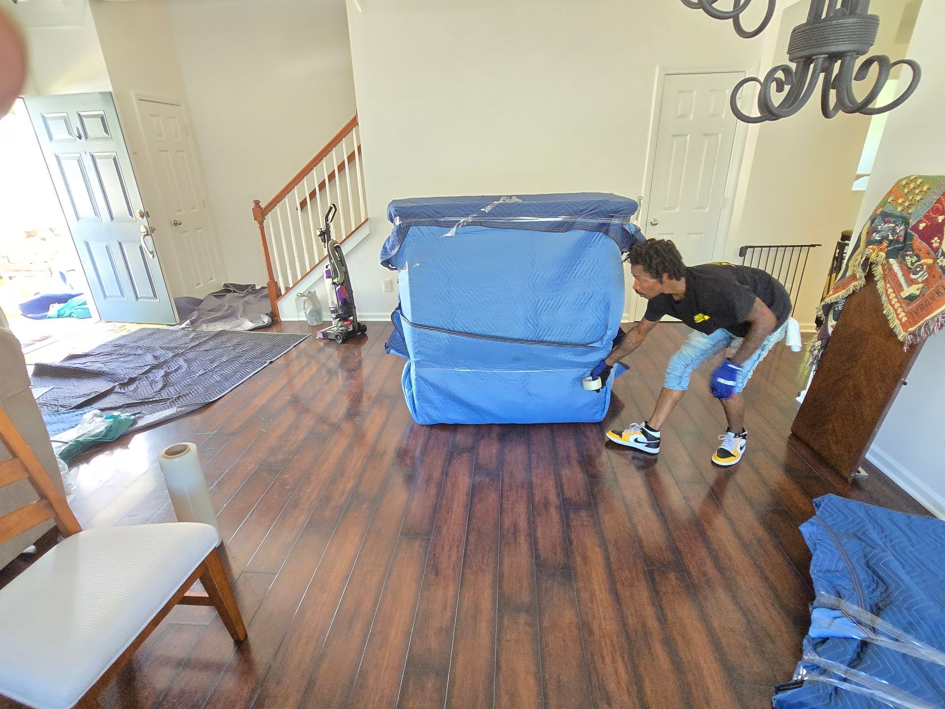 A person is moving a large, blue-wrapped piece of furniture in a brightly lit room with wood floors.