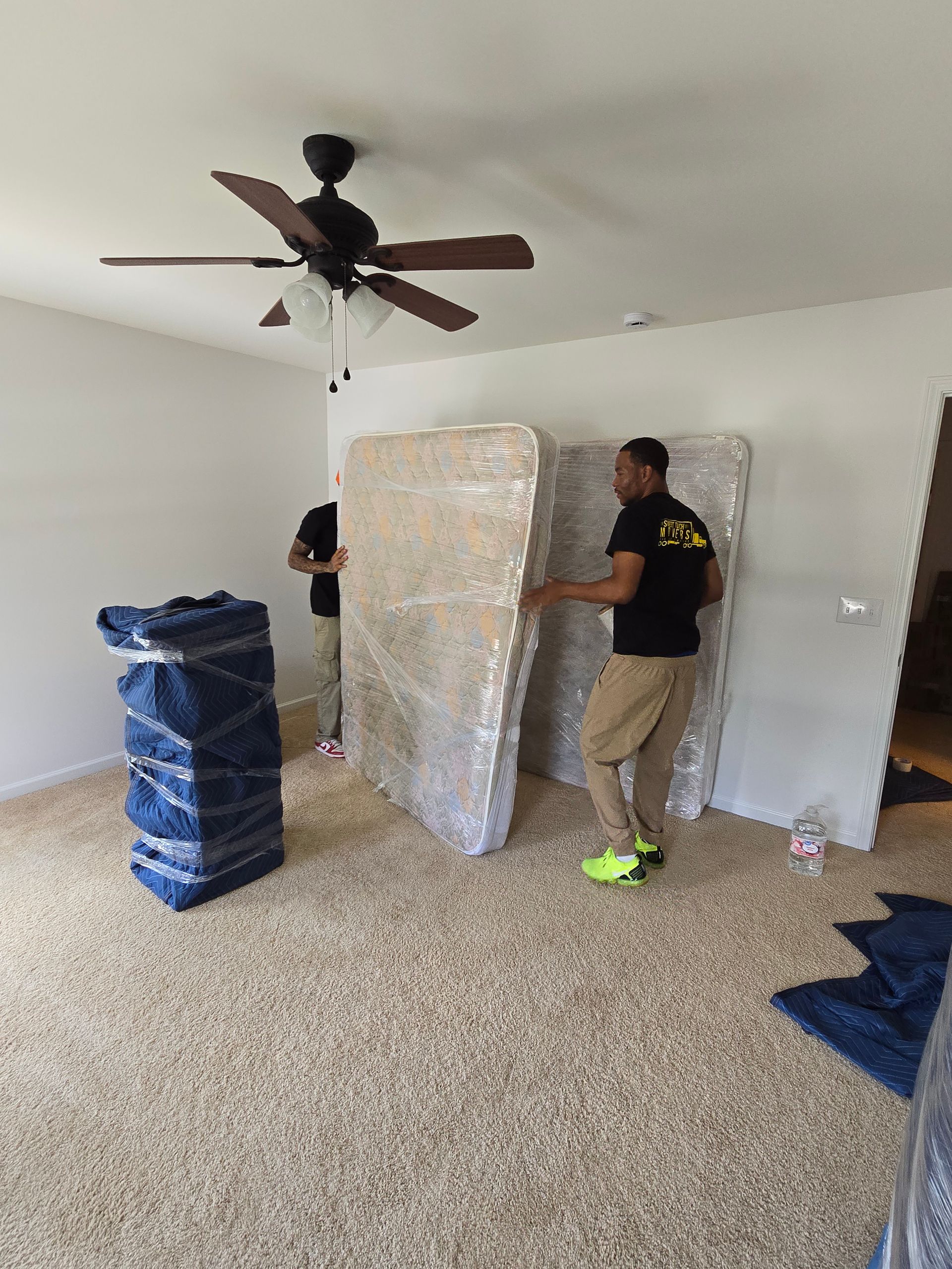 Two movers carrying wrapped mattresses into a room.  One stack of furniture wrapped in blue is on the left. Beige carpet and white walls.