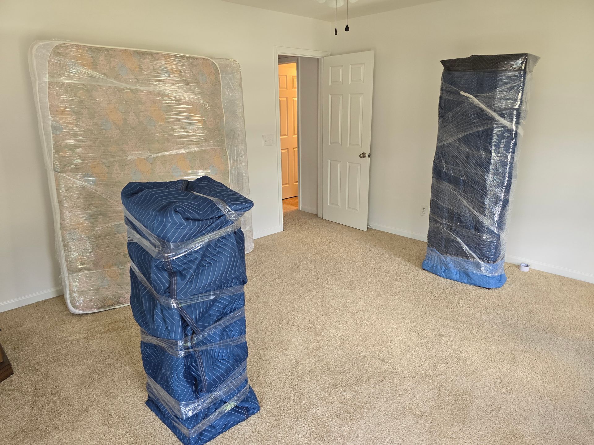 A bedroom with furniture wrapped in plastic for moving.  A mattress, two wardrobes, and stacked boxes are in the room.