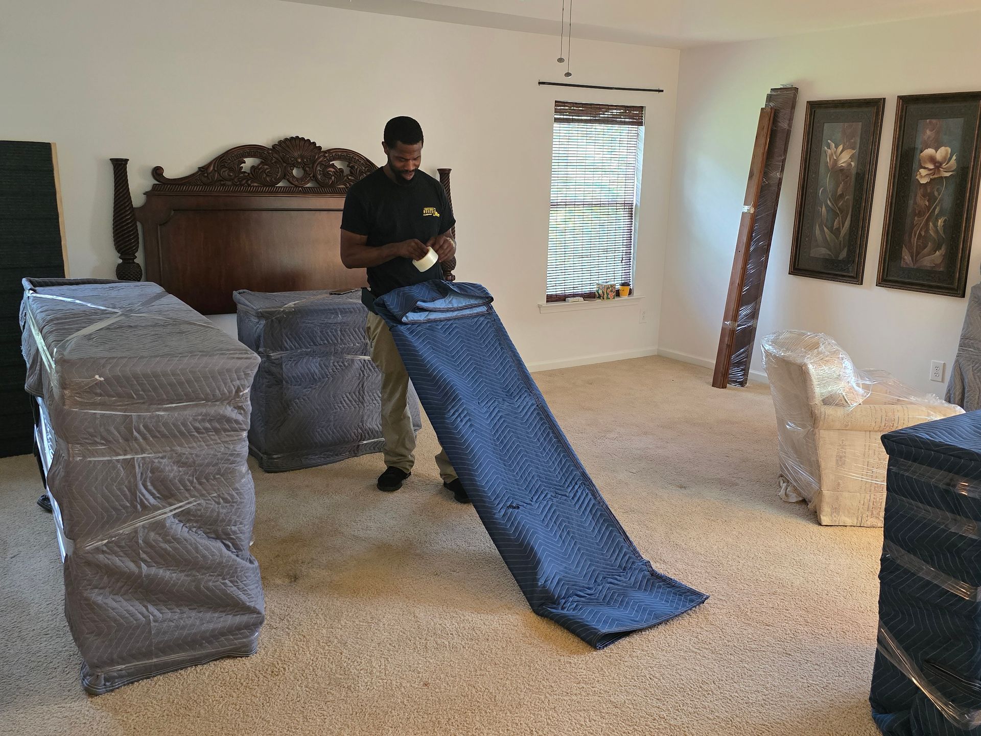 A mover prepares to load furniture into a truck, using a blue moving blanket. The room has packed furniture and art on the walls.