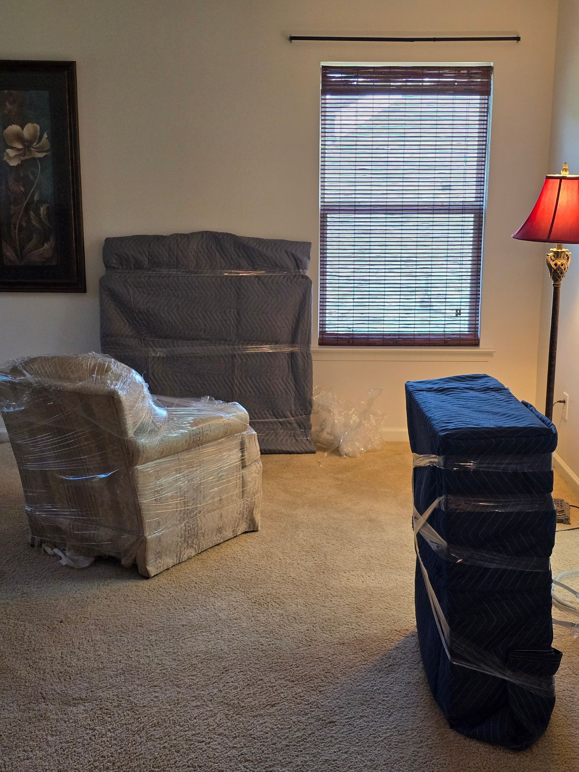 A room with furniture wrapped in plastic and moving supplies. A chair, a tall case, and a cabinet wrapped in protective coverings.