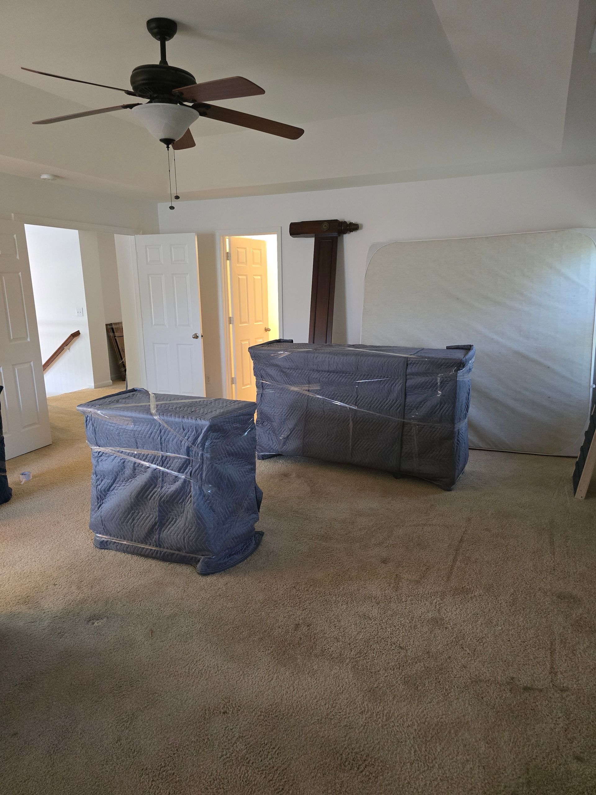 Two furniture pieces wrapped in moving blankets sit on a tan carpet in a room. A ceiling fan hangs above.