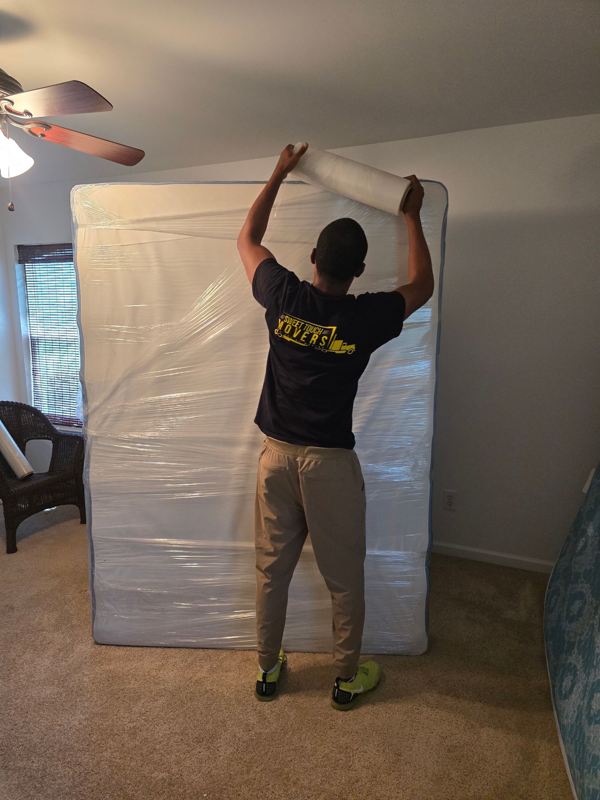 A person wrapping a large, plastic-wrapped mattress. The person is wearing khaki pants, a dark shirt, and neon green shoes in a room.