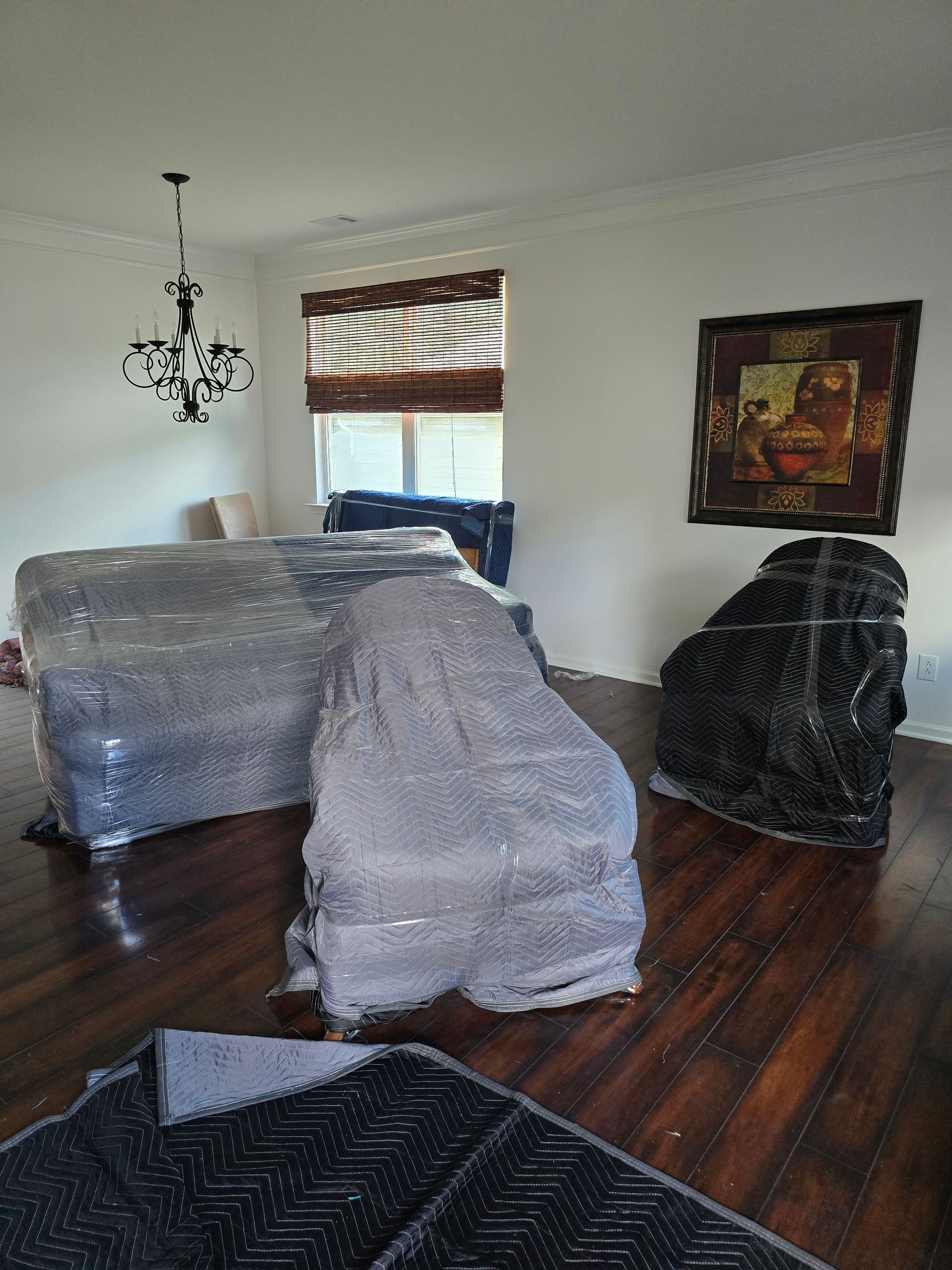 Furniture wrapped in plastic in a room with hardwood floors, ready for moving. A chandelier and artwork hang on the walls.