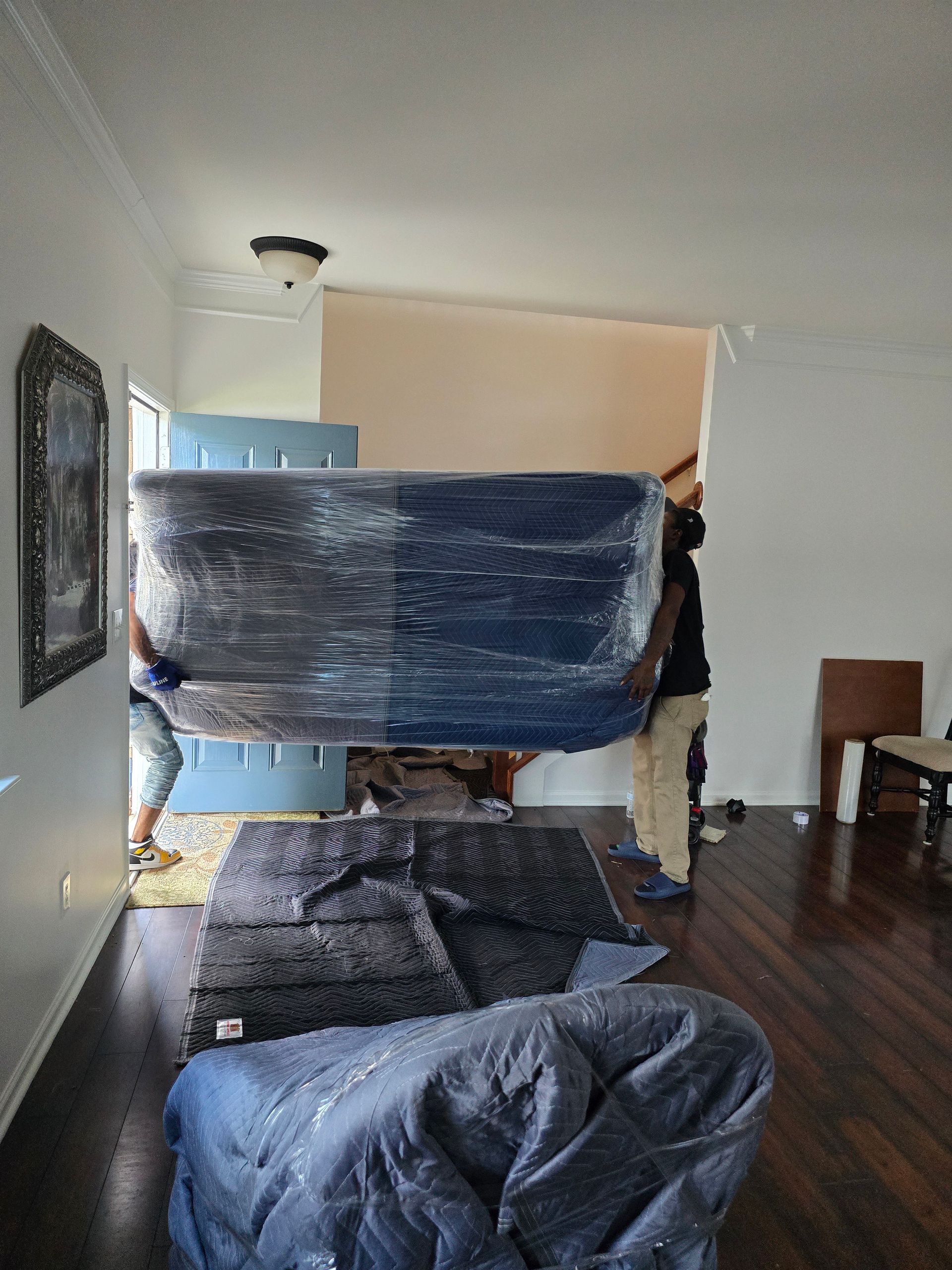 Movers carrying a large, wrapped couch through a doorway in a living room. Dark wood floors, white walls, and a rug are visible.