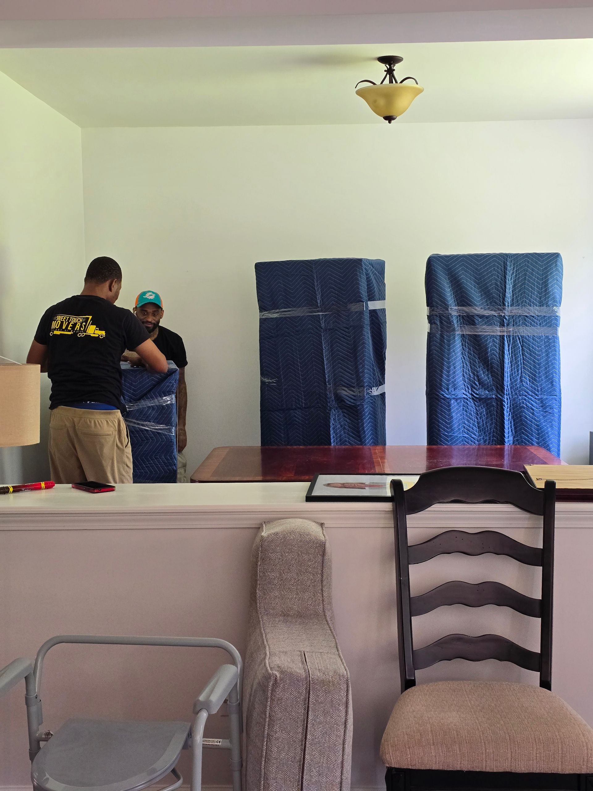 Two movers wrapping objects in blue padding inside a room. A desk, chairs, and lamp are visible.