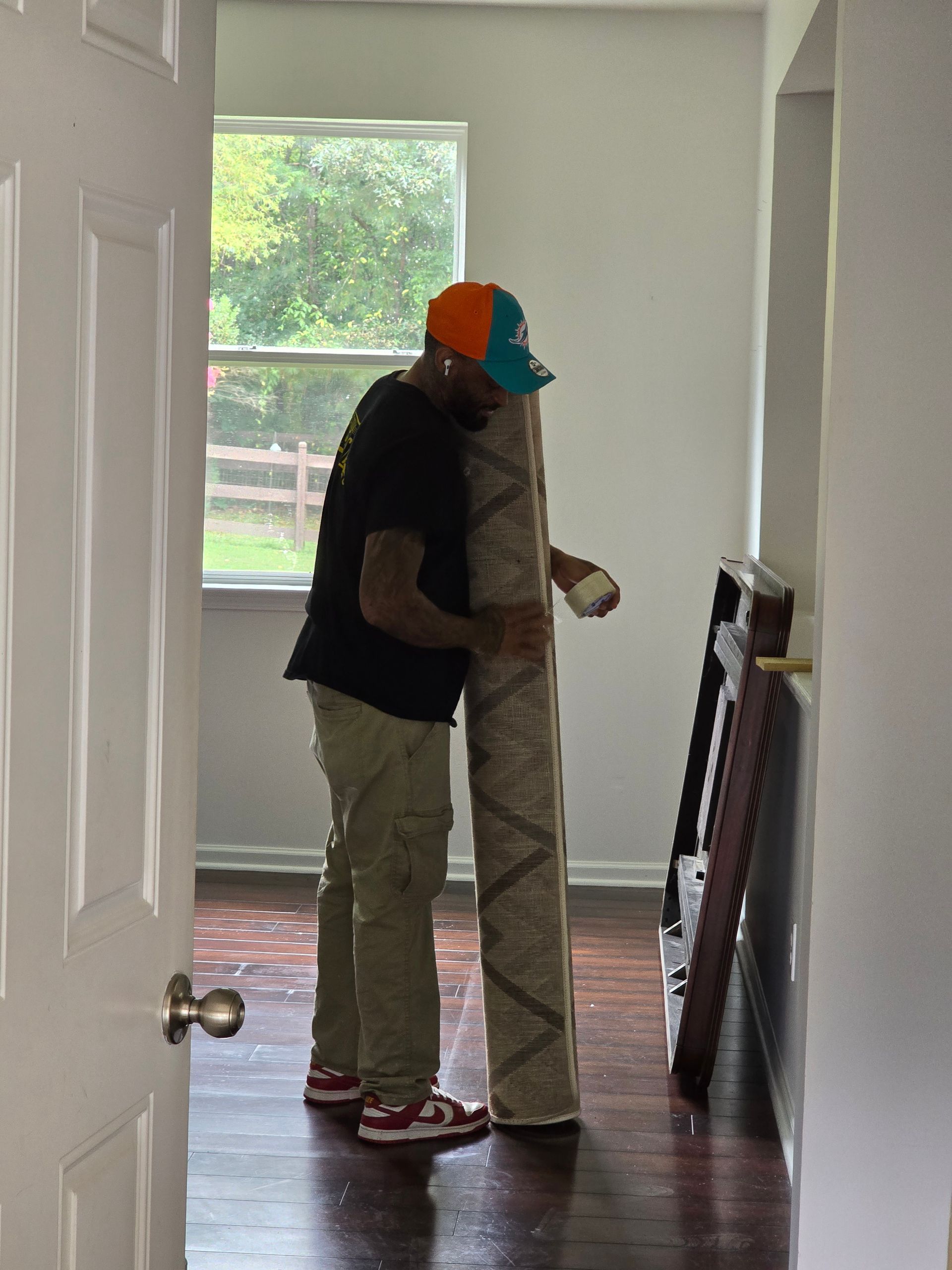 A person in a cap and cargo pants carries a rolled-up beige rug indoors; a window and doorway are visible.