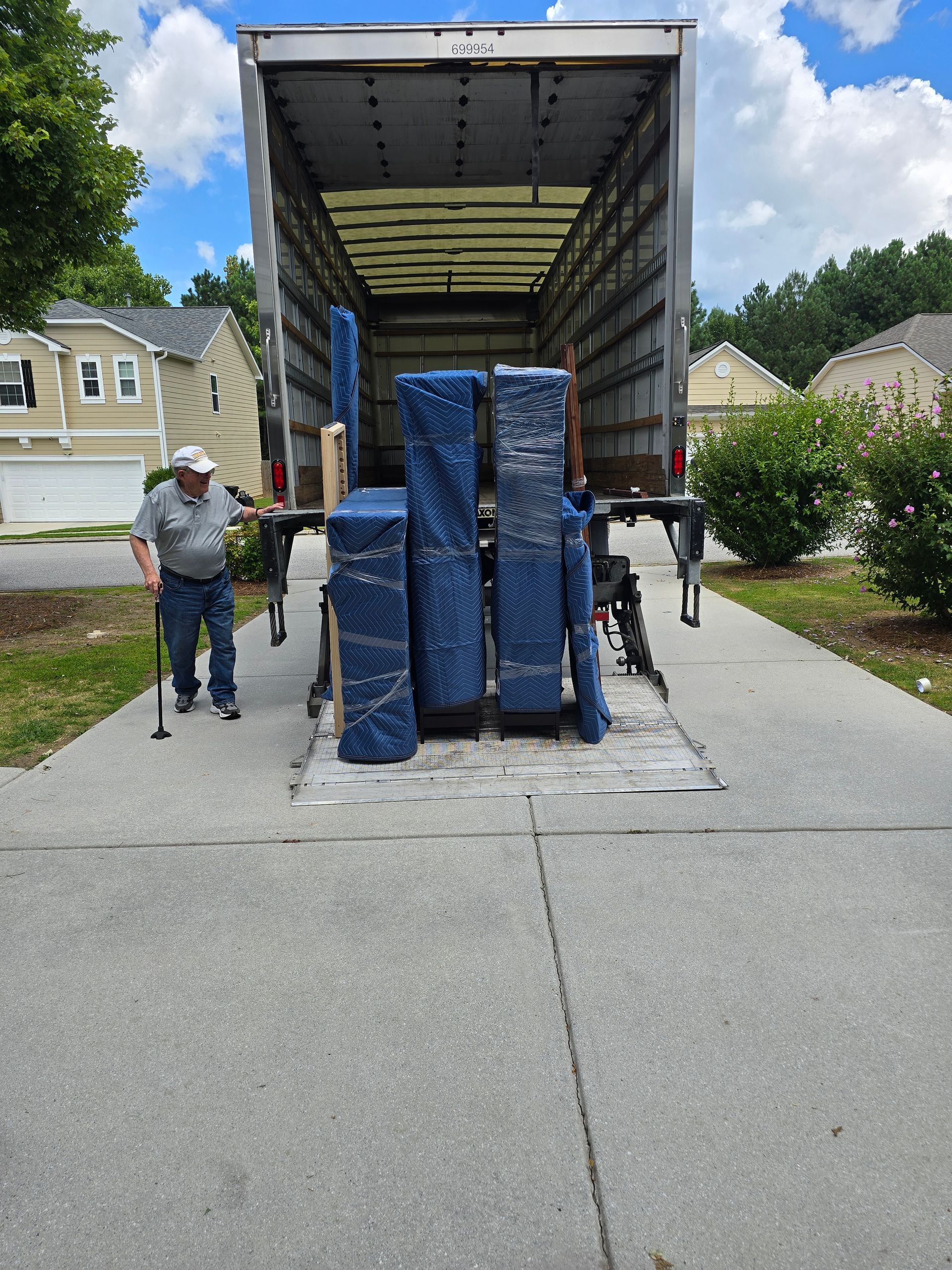 A man with a cane stands near an open moving truck filled with wrapped furniture, parked in a driveway.