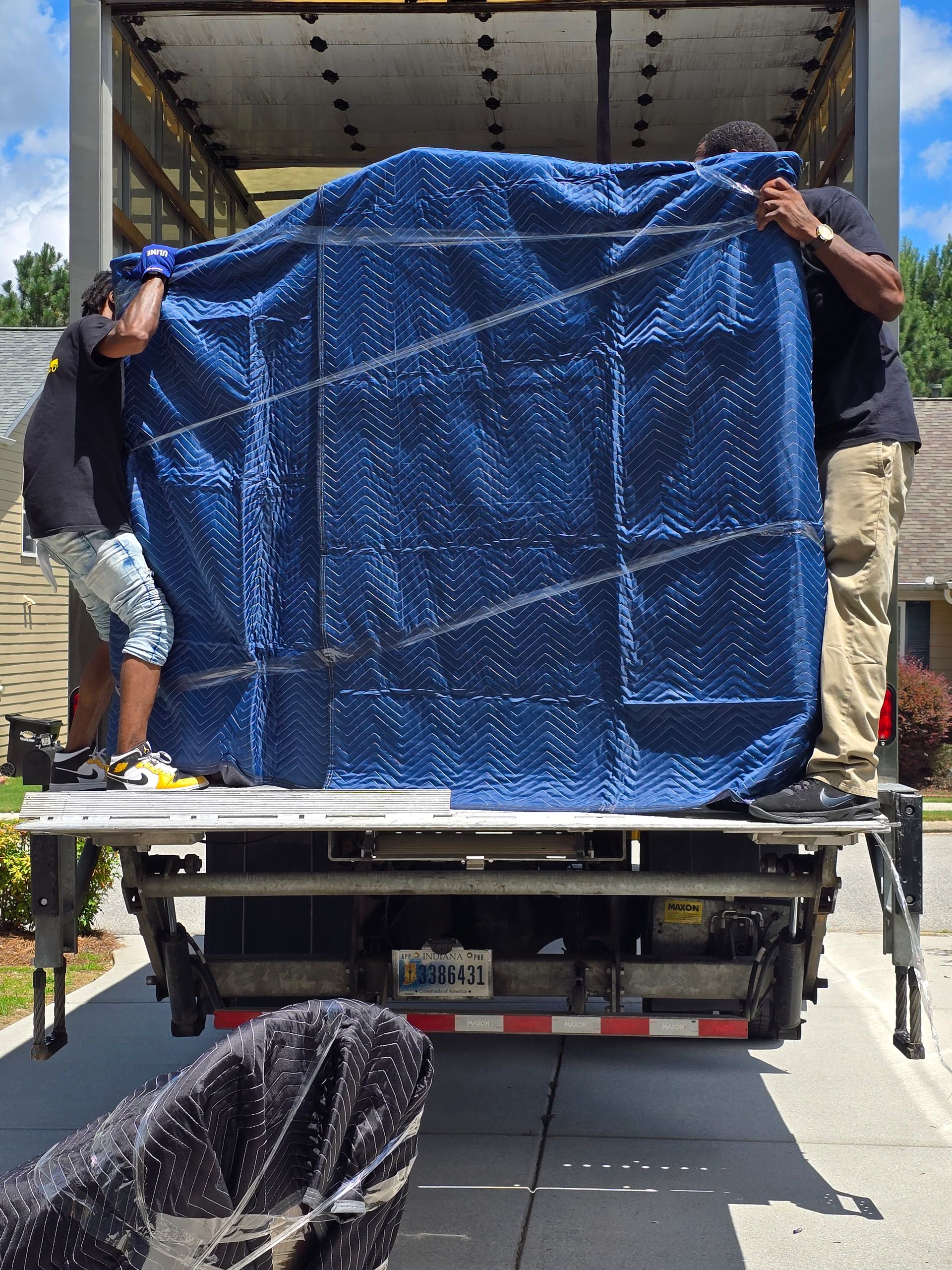 Two men loading a large, blue-covered object onto a truck from a residential driveway. Another object, wrapped in black, is visible in the foreground.
