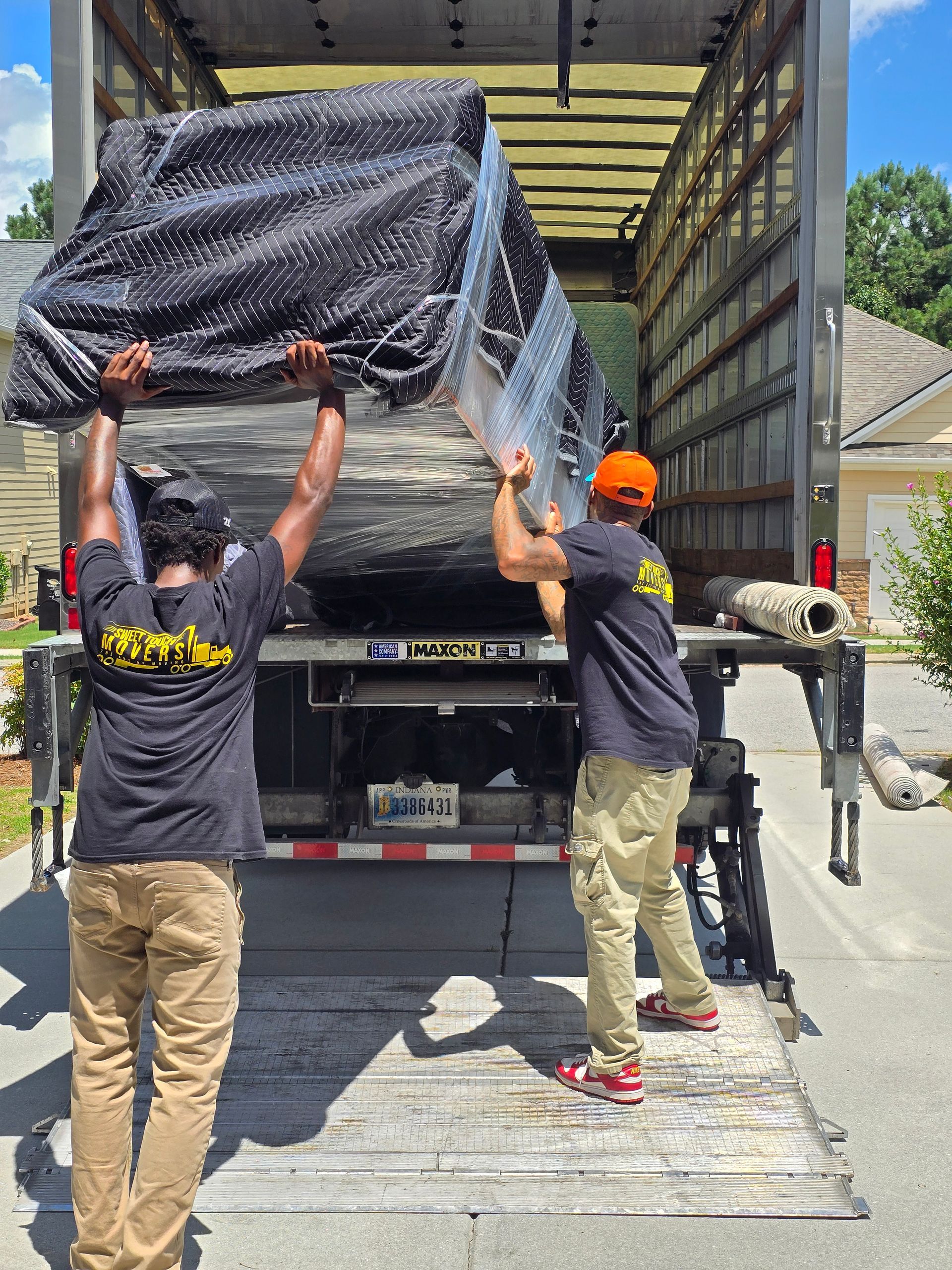 Two movers loading a wrapped couch into a truck. They are standing on the liftgate of a moving truck in front of a house.