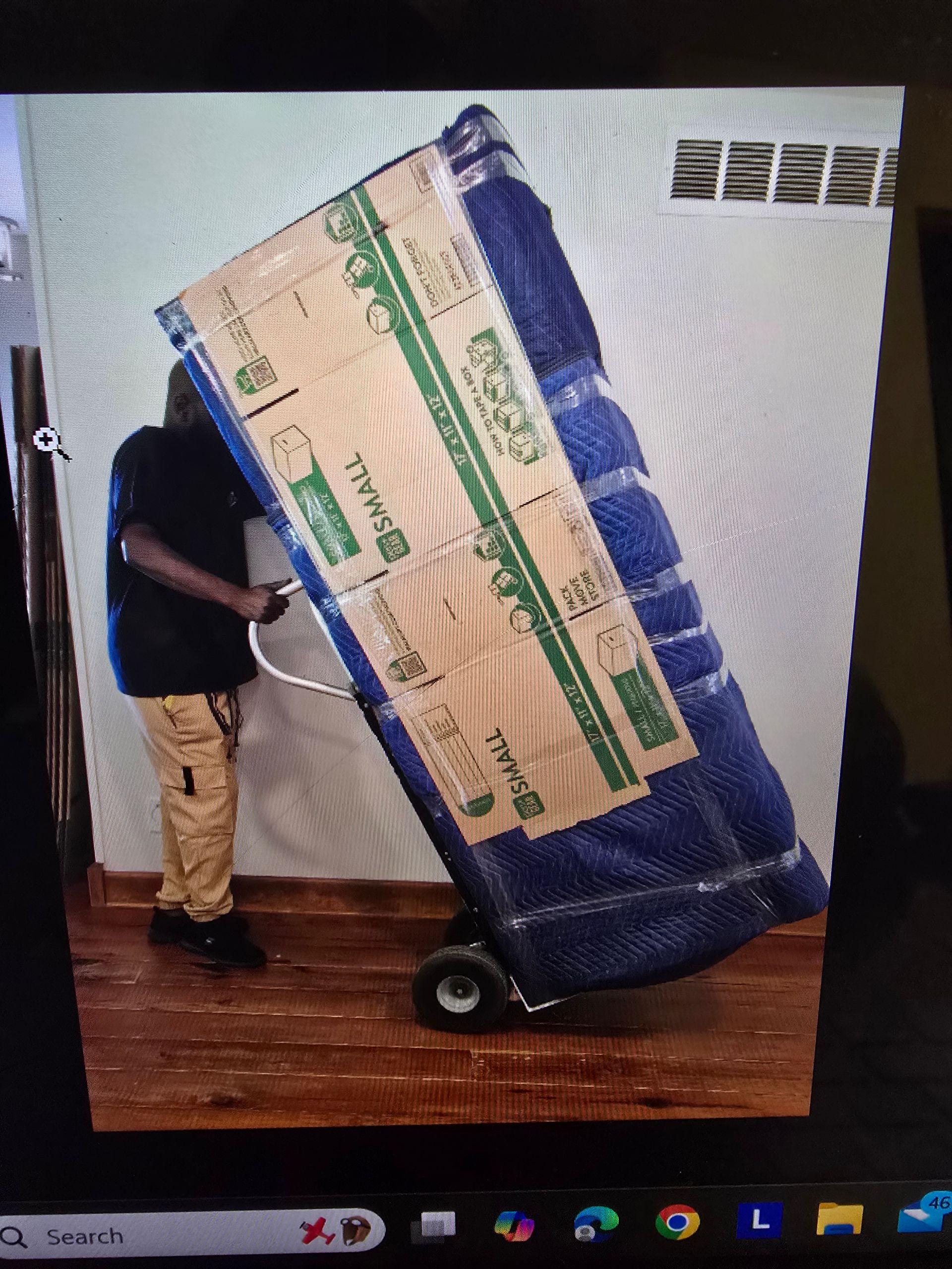 Man moving a large, wrapped item on a hand truck, likely a mattress, indoors near a wall.
