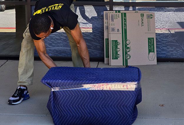 Man bending to lift a blue quilted storage bin beside stacked cardboard boxes indoors