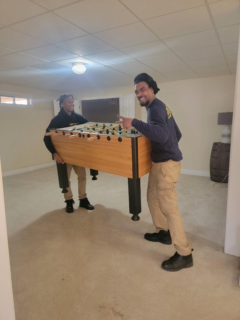 Two men are carrying a foosball table in a basement.