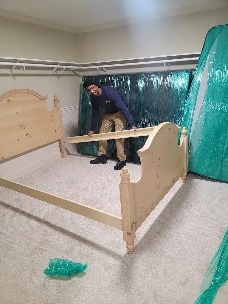 Man assembling a light-colored bed frame in a room with a patterned curtain and carpet.