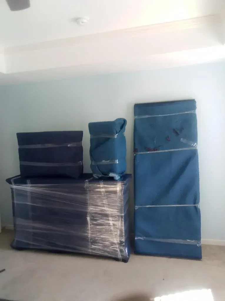 Furniture wrapped in blue moving blankets and plastic wrap, against a light blue wall in a room.