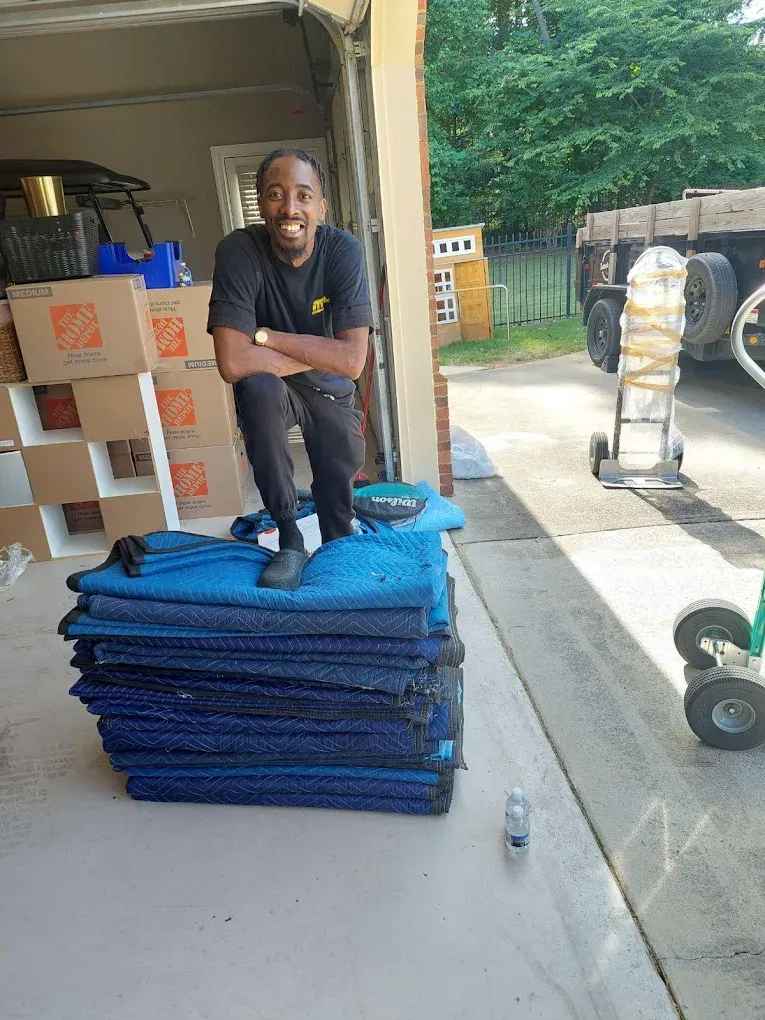 Man smiling, standing on a stack of blue moving blankets in a driveway next to a truck.