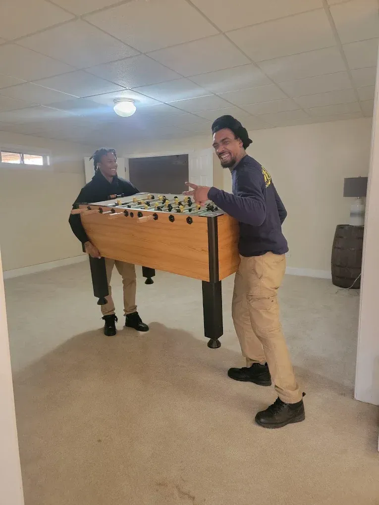 Two people carrying a foosball table in a room with beige carpet and a low ceiling.