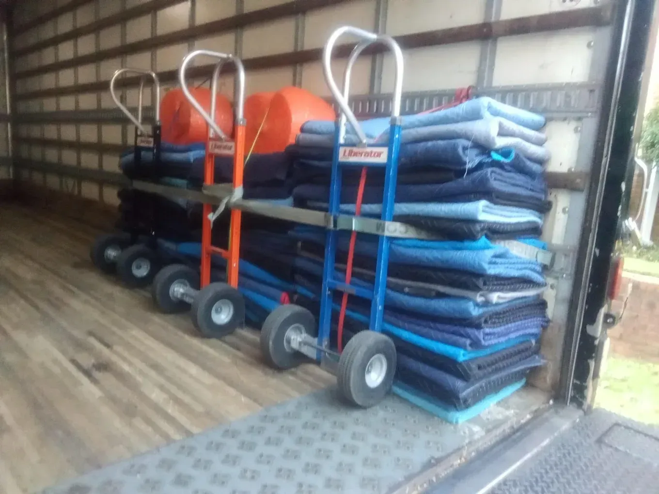 Inside a truck bed: hand trucks loaded with blue moving blankets and orange rolled items.