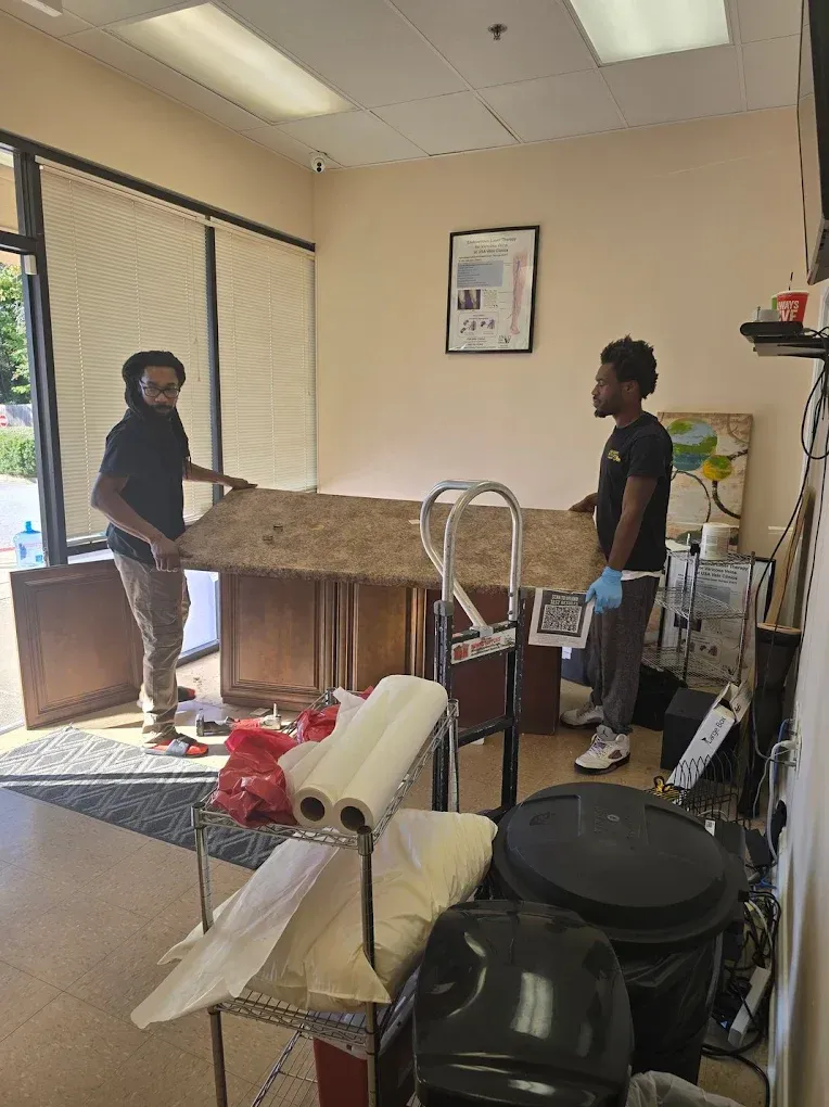 Two people lift a large wooden cabinet board inside a room. There are supplies and a trash bin in the foreground.