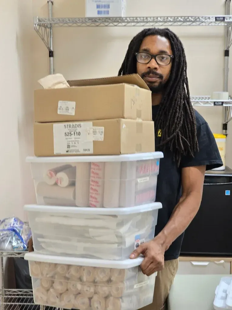Man carrying boxes and containers of supplies in a storage area; neutral expression.