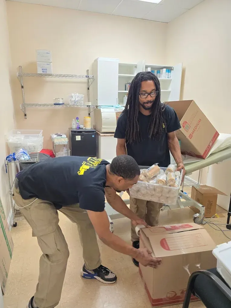 Two people moving boxes and items in an office. One holds a tray of items. Interior setting.