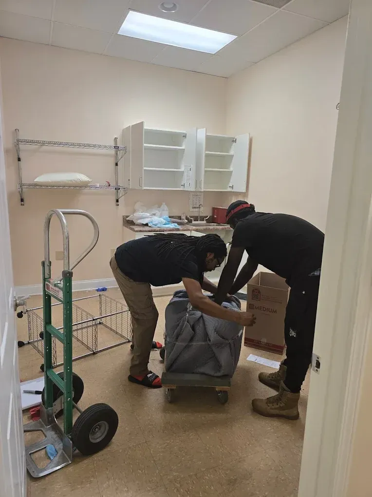 Two people moving a large, wrapped item on a dolly in a room. Shelving and cabinets are visible.