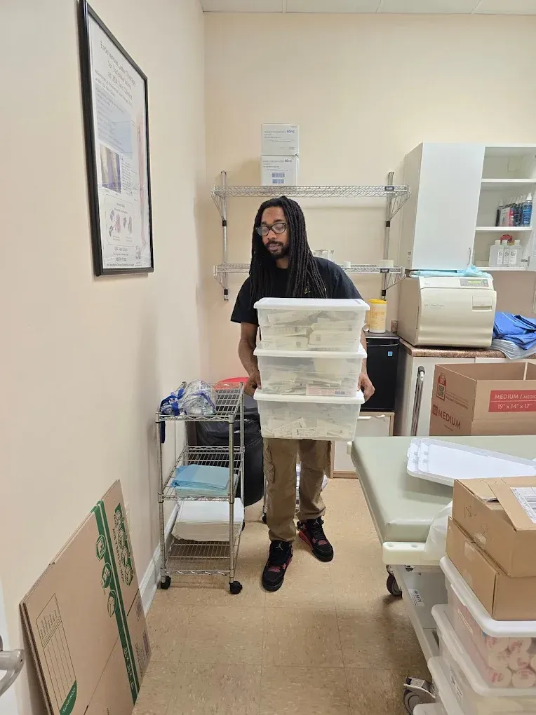 Person carrying three stacked containers in a room. Shelves, table, and boxes are also present.