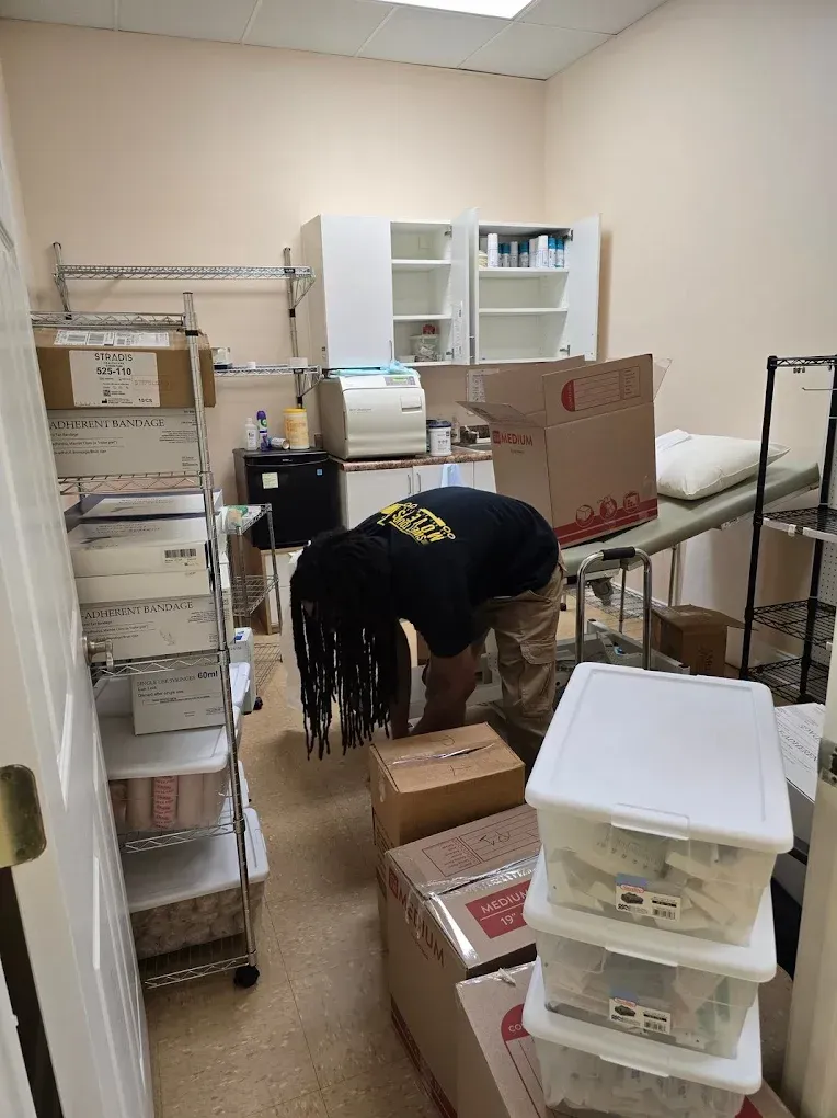 Person in a cluttered room, looking into a box. Shelves, boxes, and medical equipment are present.