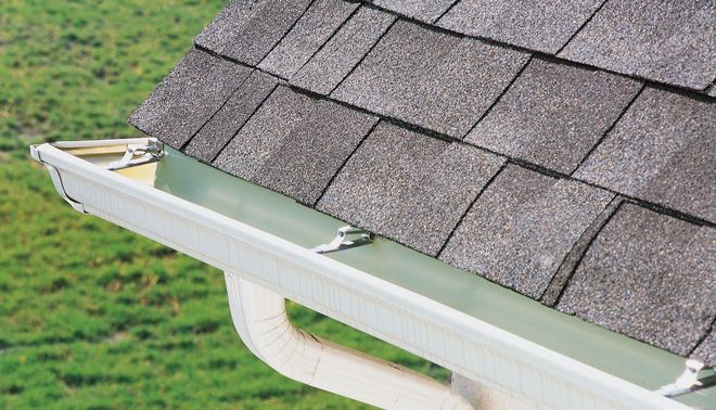 Close-up of a roof gutter collecting rainwater beside gray asphalt shingles.