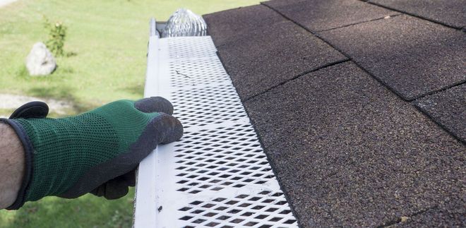 Gloved hand installing a white roof gutter guard along shingle roofing edge