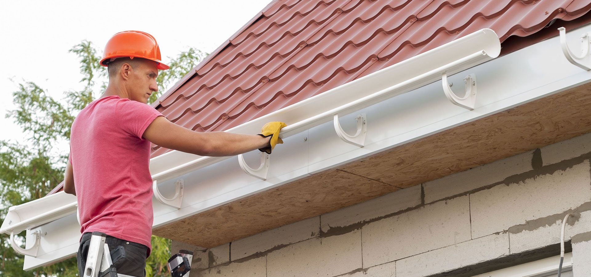 Worker in orange hard hat installing a white gutter on a house roofline