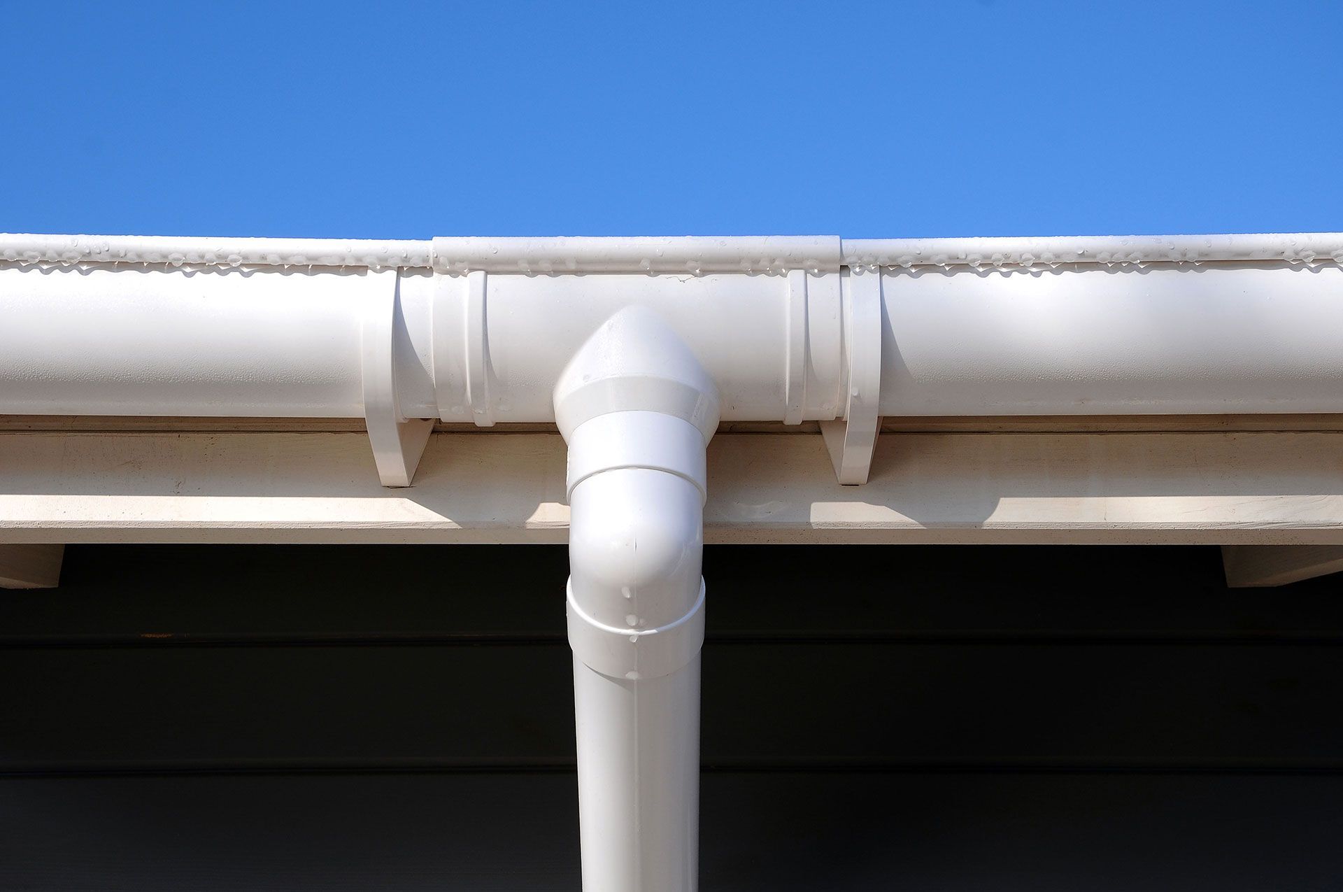 White PVC downspout connected to a gutter against a blue sky and roofline
