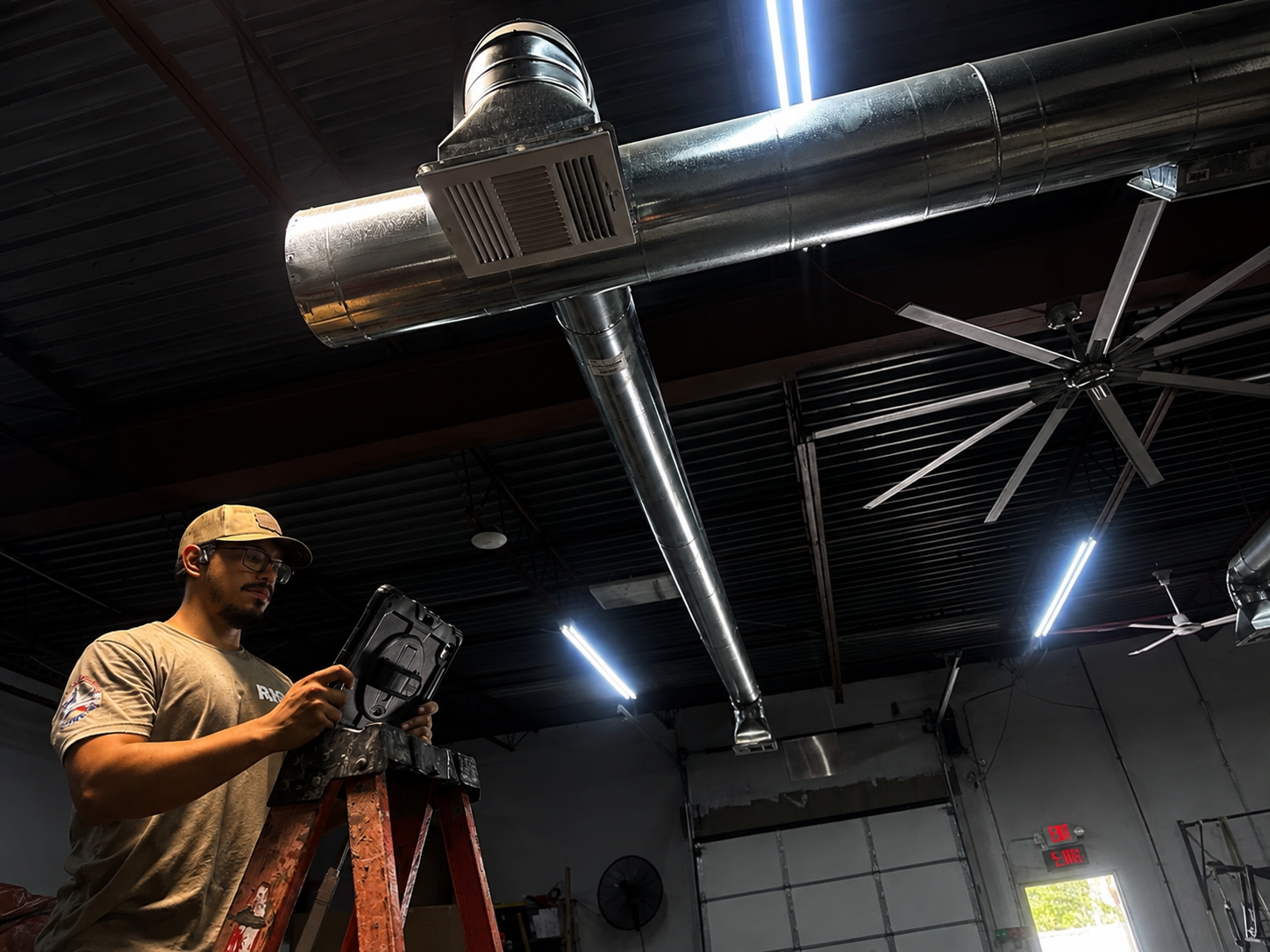 Worker on a ladder inspecting large silver ductwork in an industrial ceiling room