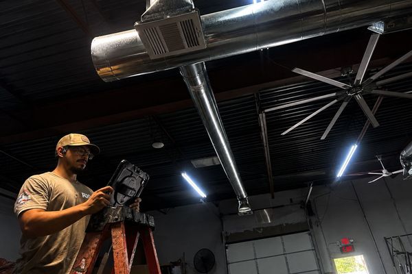 Man in a cap adjusting a stage light in a dark industrial room with ceiling ducts and fans