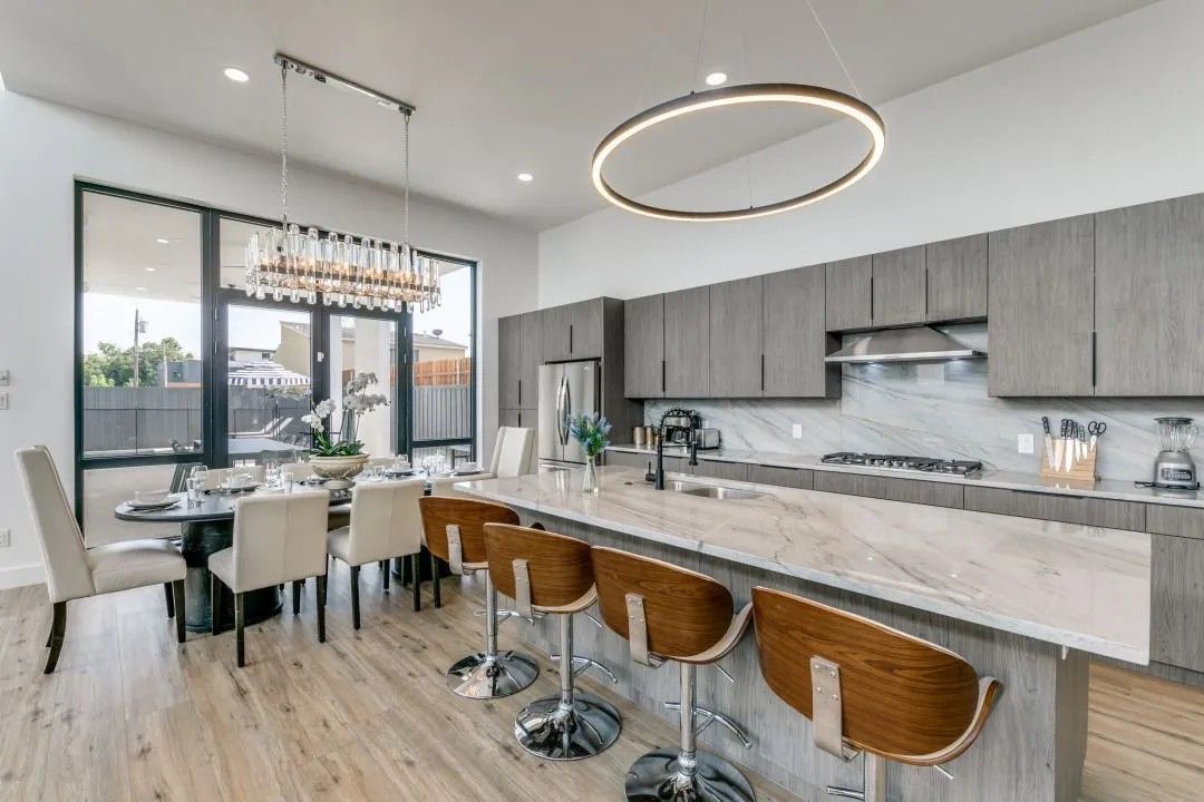 Modern kitchen with a large island, gray cabinets, and a dining table near large windows.  Wooden floors and stylish light fixtures.