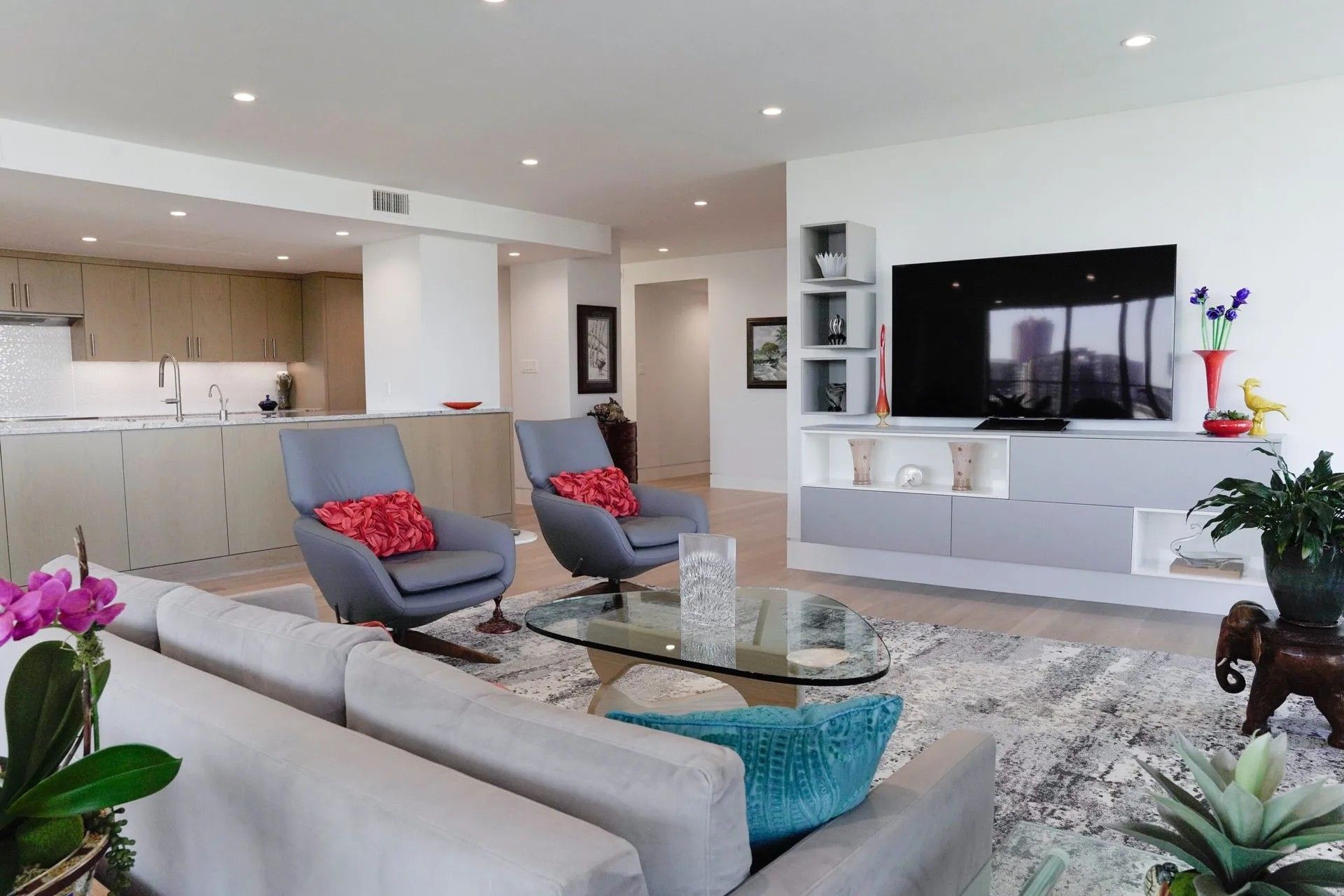 Modern living room with gray sofa, two armchairs, glass coffee table, and a large TV. The space also includes a kitchen, shelves, and plants.